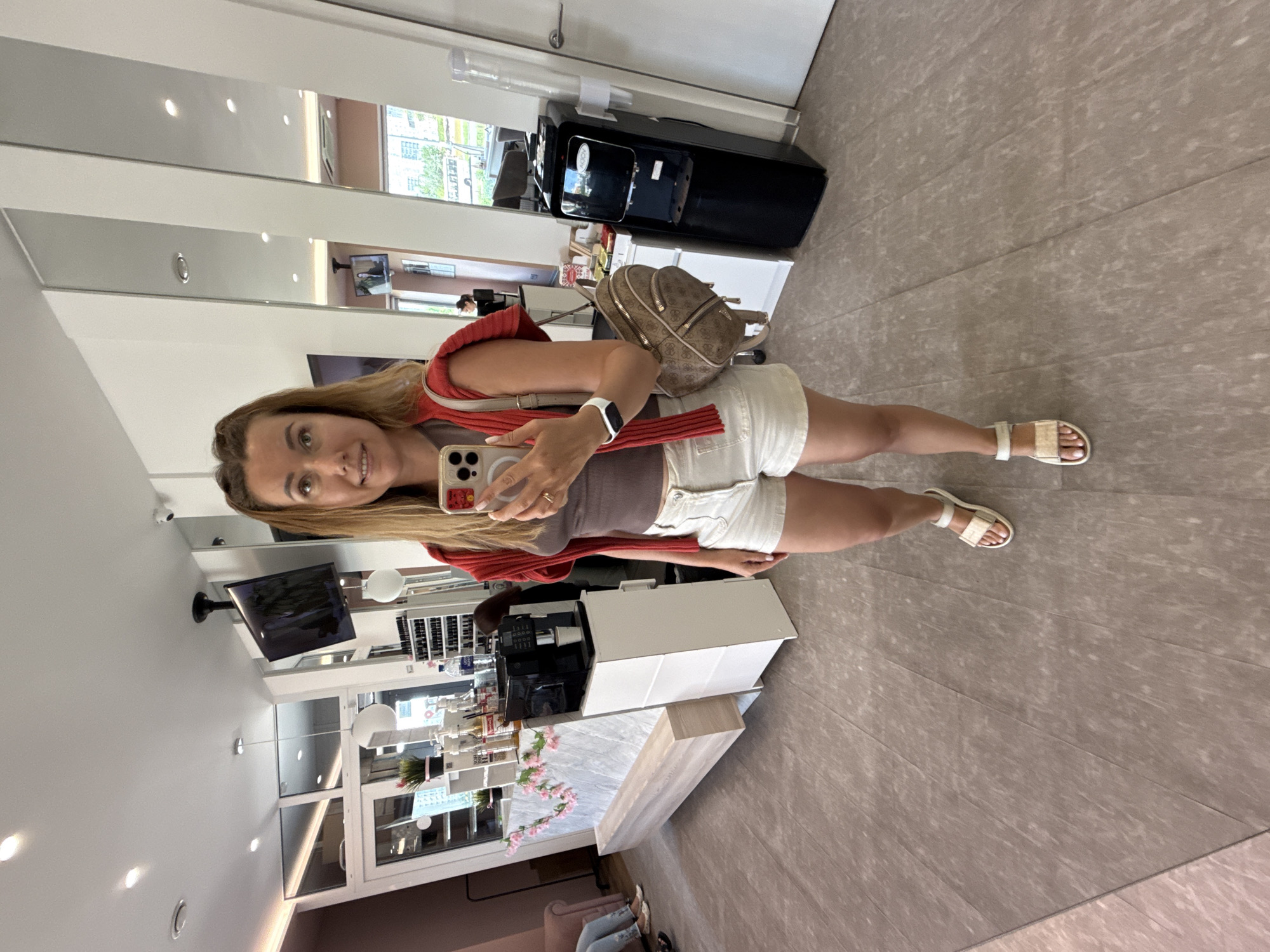 A woman with blonde hair pulled back and braided is taking a mirror selfie in an indoor establishment. She is wearing a brown tank top, white shorts, a red knit cardigan draped over her shoulders, and white sandals. She has a beige backpack with a patterned logo on her right shoulder. In her right hand, she holds a smartphone, which she is using to take the photo. Her left wrist has a white smartwatch.
The environment appears to be a modern retail or service space, possibly a salon or cafe, with polished light-colored flooring. To the left, there is a counter with a marble top decorated with pink artificial flowers. A coffee machine is visible on the counter, with a white cup next to it. Behind the counter, shelves hold bottles and products. A water cooler is present on the right side of the image. Mirrors line the back wall, reflecting the interior. In the background, a television screen is mounted on the ceiling, displaying an image. Other patrons are partially visible in the background, with one person seated on a pink sofa on the far left. The overall lighting is bright and artificial.