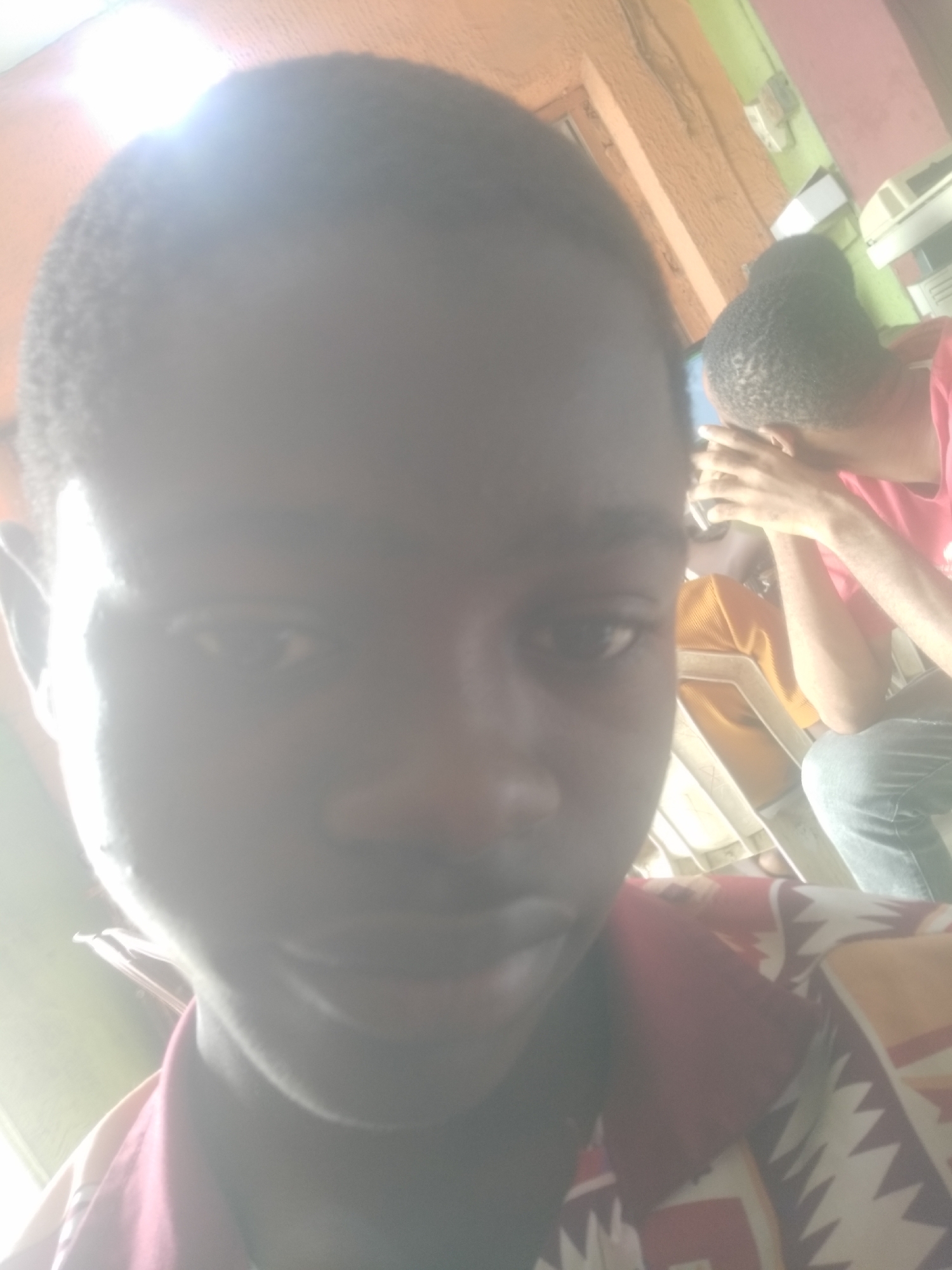 The image is a selfie of a young man with dark skin and short hair. He is wearing a patterned shirt with a maroon collar. In the background, another person is seated and wearing a red shirt and jeans. The environment appears to be indoors with walls painted in multiple colors. The location is likely in Shangisha, Nigeria.