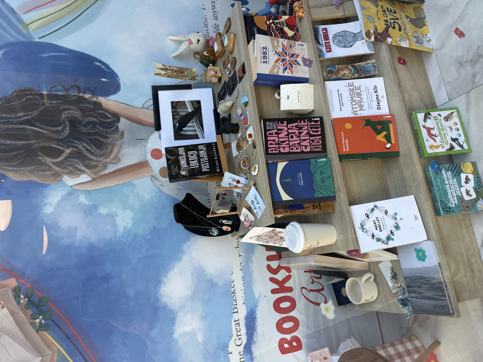 Here's a concise description of the image:
**Scene:** An outdoor market stall likely in Budva, Montenegro, displaying books and handcrafted items. The stall is set up on a light-colored surface (possibly a large print) that includes the word "Booksy."
**Objects:** Books are the dominant objects, with visible titles and authors including "Nepodnošljiva lakoća postojanja" by Milan Kundera, "1983" by Miljenko Jergović, "Arja" by Hristina Radočić Andrić, "Atomske navike" by James Clear, "Mary Shelley Frankenstein" and "Cudesna Crna Gora." Other objects include jewelry, small decorative items (a rabbit doll, small shoes), a white coffee cup with a lid, a mug, framed photos, and wooden clothes pegs.
**Environment:** The setting appears to be outdoors in daylight. The underlying "Booksy" print suggests a connection to literature or art. The overall impression is that of a curated display intended to attract attention, possibly part of a local market or fair.