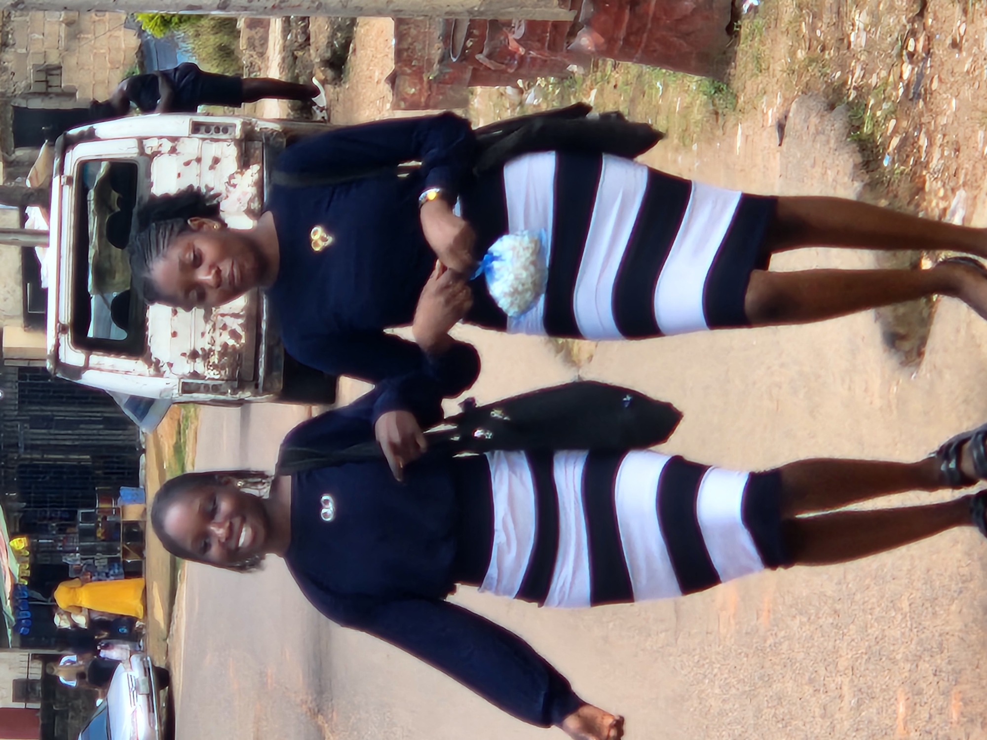 Two young women are walking on a dirt road in Jos, Nigeria, in front of a dilapidated white van. The woman on the left wears a dark blue long-sleeved shirt and a black and white striped skirt. She carries a black tote bag over her left shoulder. The woman on the right also wears a dark blue long-sleeved shirt and a black and white striped skirt. She holds a small white bag in her right hand and carries a black tote bag over her left shoulder. The background shows a street scene with buildings, a car, and several barrels. The overall environment appears to be urban and somewhat run-down.