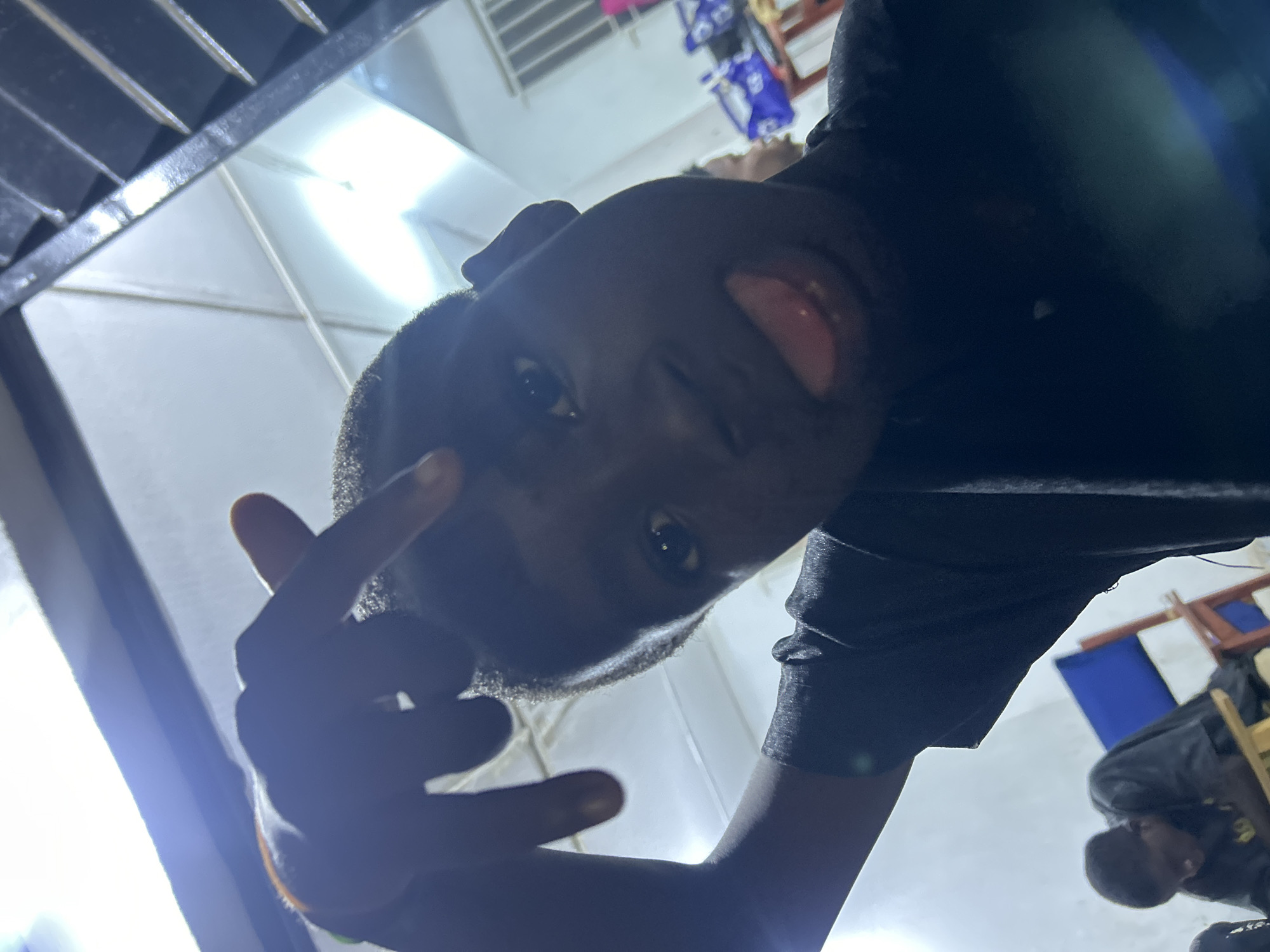The image shows a person of dark complexion making a gesture with their fingers and sticking out their tongue, captured from a low angle looking upwards. The person appears to be wearing a dark-colored shirt. The background includes a light-colored ceiling and a window with horizontal bars. Further in the background, other people and objects are visible but less distinct due to the focus being on the individual in the foreground. The location appears to be indoors, and the specific city and further details of the environment are not discernible from the image.