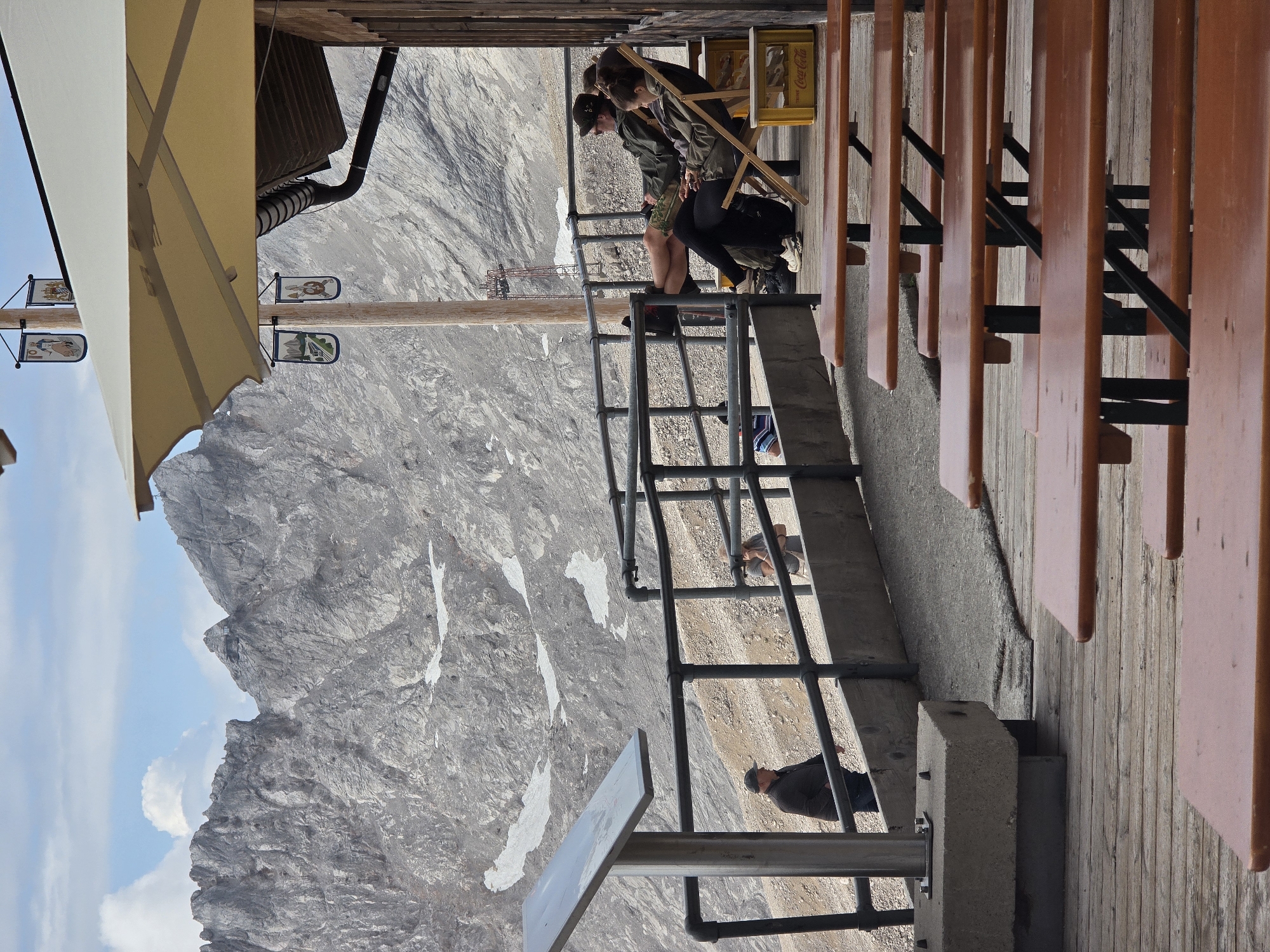 This image depicts a mountain restaurant with a view of Zugspitze mountain, Germany's highest peak, near Garmisch-Partenkirchen. In the foreground, several empty wooden picnic tables are arranged on a wooden deck. Further back, a metal railing separates the deck from a slope. A man in a dark jacket and cap stands near a metal information sign. Behind the railing, two people are seated on wooden chairs, with one person in the foreground looking at a phone and the other in the background with their legs crossed. A yellow Coca-Cola crate is visible next to one of the chairs. The background is dominated by the rugged, rocky slopes of the mountains, with patches of snow. A cable car structure is visible on the mountainside. The sky is partly cloudy.
