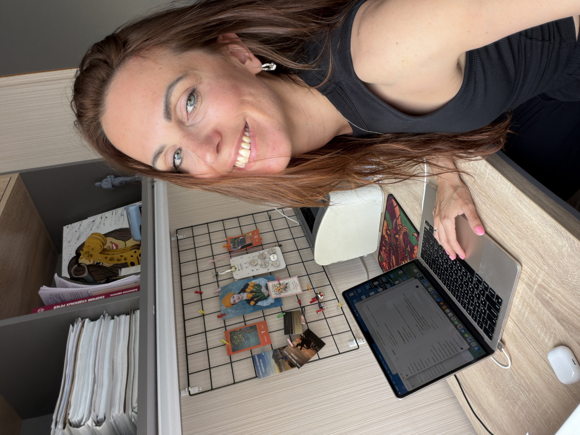 A woman with long, reddish-brown hair smiles at the camera while sitting at a desk. She is wearing a black tank top and has bright blue eyes. She is typing on a laptop, and a wireless earbud case sits beside it. Behind her, a black wire grid is decorated with various photos and postcards. The desk is made of light-colored wood. Above the desk, a shelf holds a stack of papers and some books. The scene appears to be in a home office or study. There is no specific indication of the location in Birobidzhan, Russia.