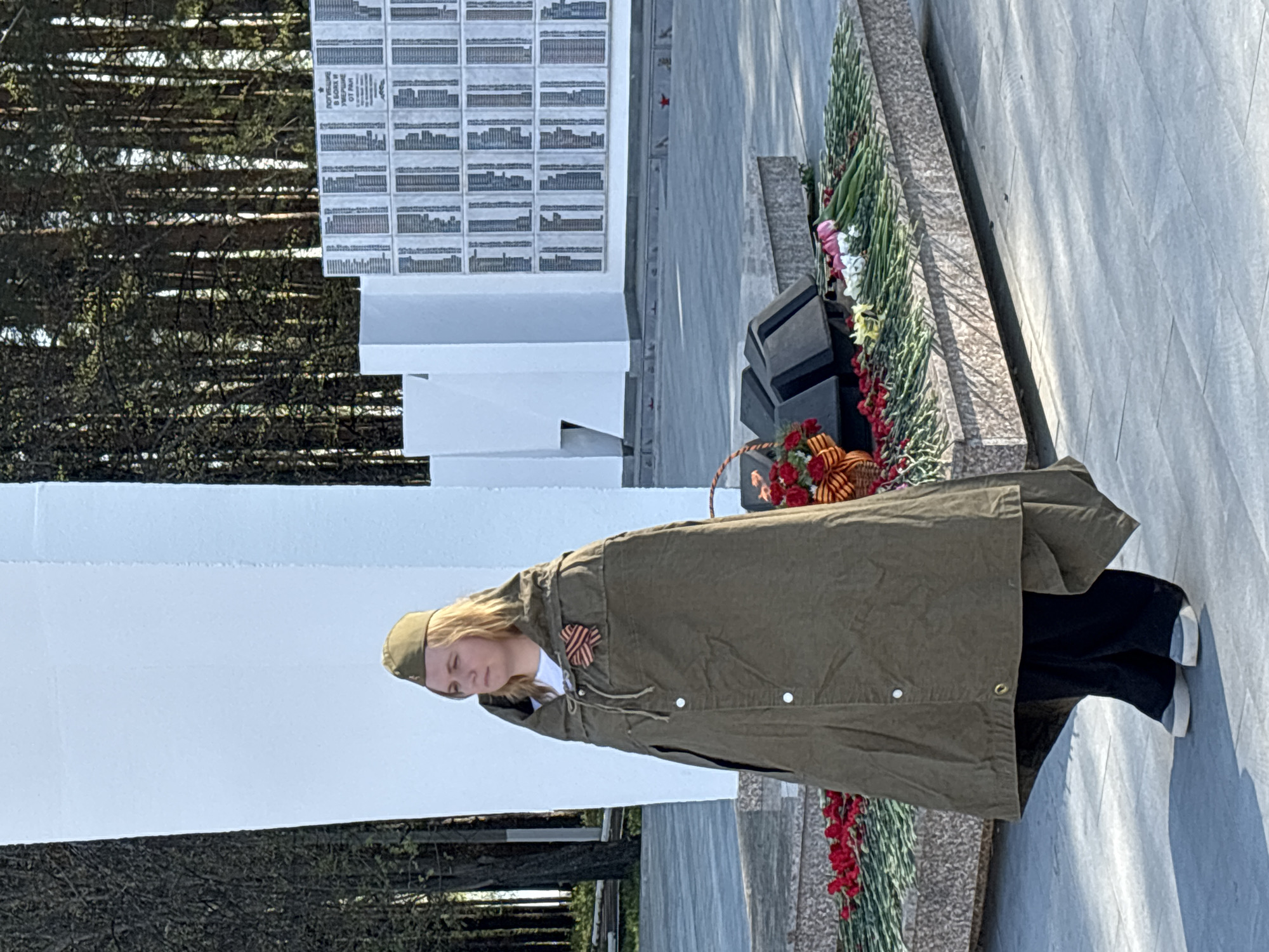 A woman in a green coat and cap, adorned with a St. George ribbon, stands near a memorial. She is near a basket of red flowers and ribbons. The memorial consists of stacked black blocks, a granite base with flower arrangements, and a white wall covered with memorial plaques. The location is Chaikovsky, Russia. There are evergreen trees in the background.