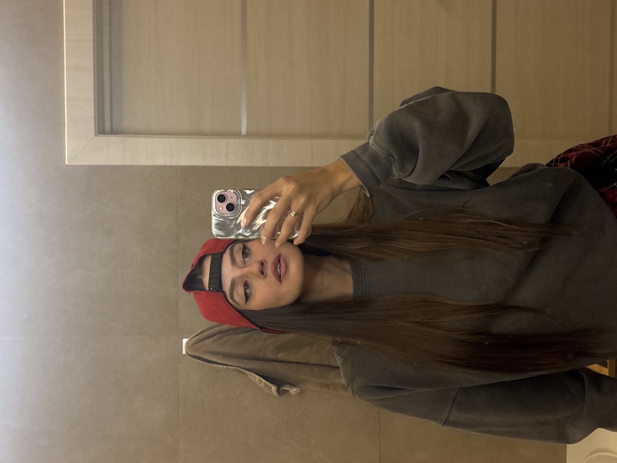 A young woman is depicted taking a mirror selfie indoors. The image is oriented vertically, showing her reflection from the chest up. She holds a smartphone, likely an iPhone with dual cameras and a silver, textured case, in her right hand, which is raised to capture her image. A gold ring is visible on her right ring finger.
She wears a red baseball cap reversed on her head, with the black adjustable strap visible at the back. Her long, straight dark brown hair flows over her left shoulder. She is dressed in a loose-fitting, charcoal gray long-sleeved sweatshirt. Her facial expression is neutral, with her eyes looking directly towards the phone's lens. She has distinct winged eyeliner and a subtle lip color.
The background of the selfie shows a simple, neutral-toned interior, possibly tiled walls or a smooth surface, with a light-colored wooden frame or cabinet structure visible in the upper portion of the reflection. A piece of red and black plaid fabric or clothing is partially visible on the far right side of the image, behind her. The setting appears to be an indoor space in Krasnoyarsk, Russia, with soft, even lighting. There is no visible text in the image.