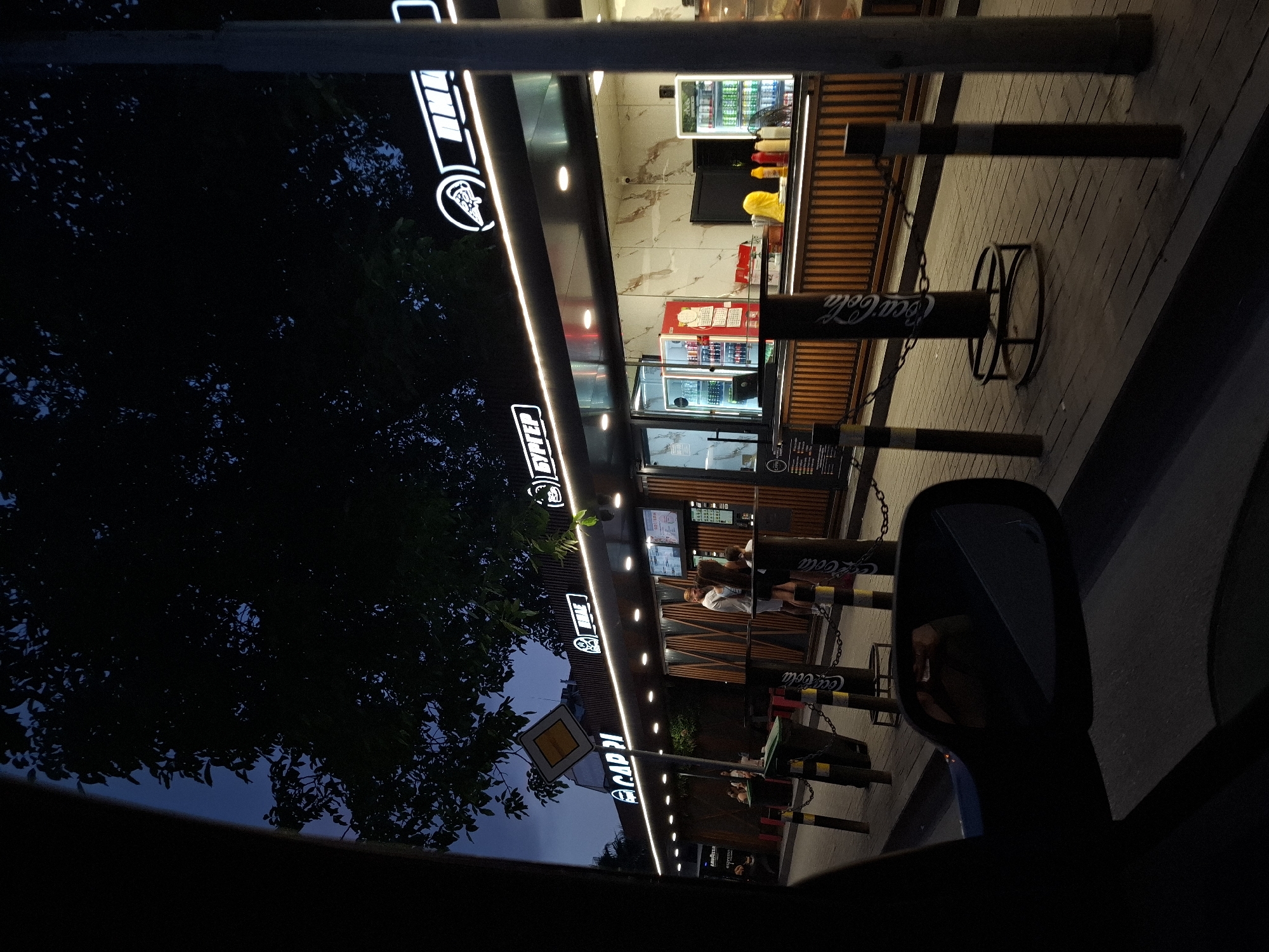The image shows a street-level view of a food establishment at dusk or night. The establishment has a modern facade with illuminated signage. The signage includes text in what appears to be Cyrillic script, with "БУРГЕР" (Burger) visible. There are also logos and names such as "CAP 21" and "LAVAZZA ESPRESSO". The food stall has a counter where staff are serving customers. Inside, refrigerators display drinks, and there are condiments and food preparation areas. People are standing in line, ordering, or waiting for food. The pavement in front of the establishment is paved with bricks. Various bollards with chains and Coca-Cola branding are present, acting as a barrier. A car's side mirror is visible in the foreground, suggesting the image was taken from inside a vehicle. Trees with dark foliage are silhouetted against the twilight sky. The overall scene depicts a busy outdoor dining or takeaway food service area in an urban setting.