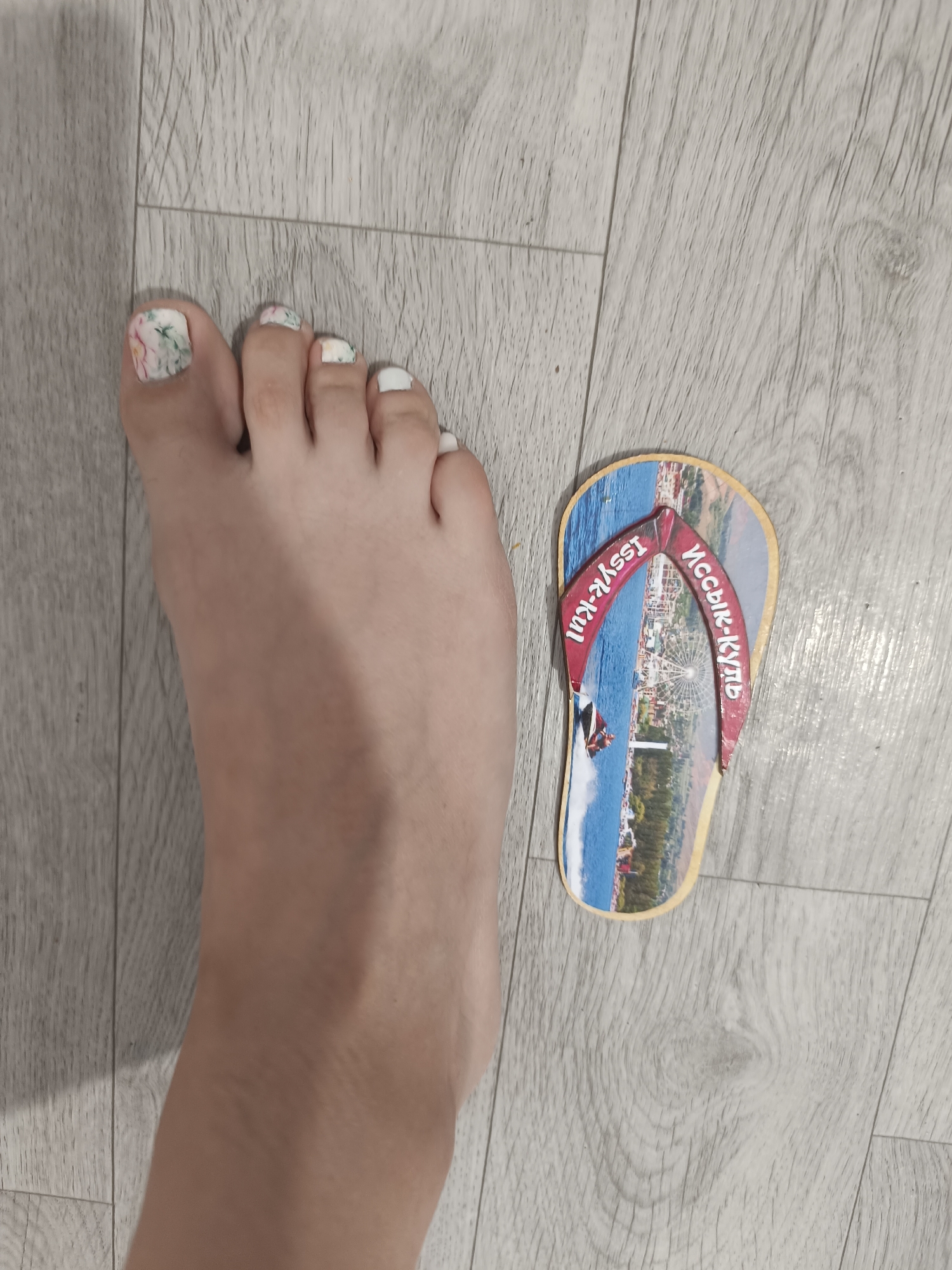 A left foot with floral patterned nail polish on some toes and white polish on others rests on a light-colored wooden floor. Next to the foot is a novelty flip-flop shaped magnet depicting a waterfront scene with a Ferris wheel. Text in both English ("Issyk-Kul") and Cyrillic ("Ыссык-Куль") is present on the red strap of the magnet. The image does not show the city administration of Taraz, Kazakhstan.