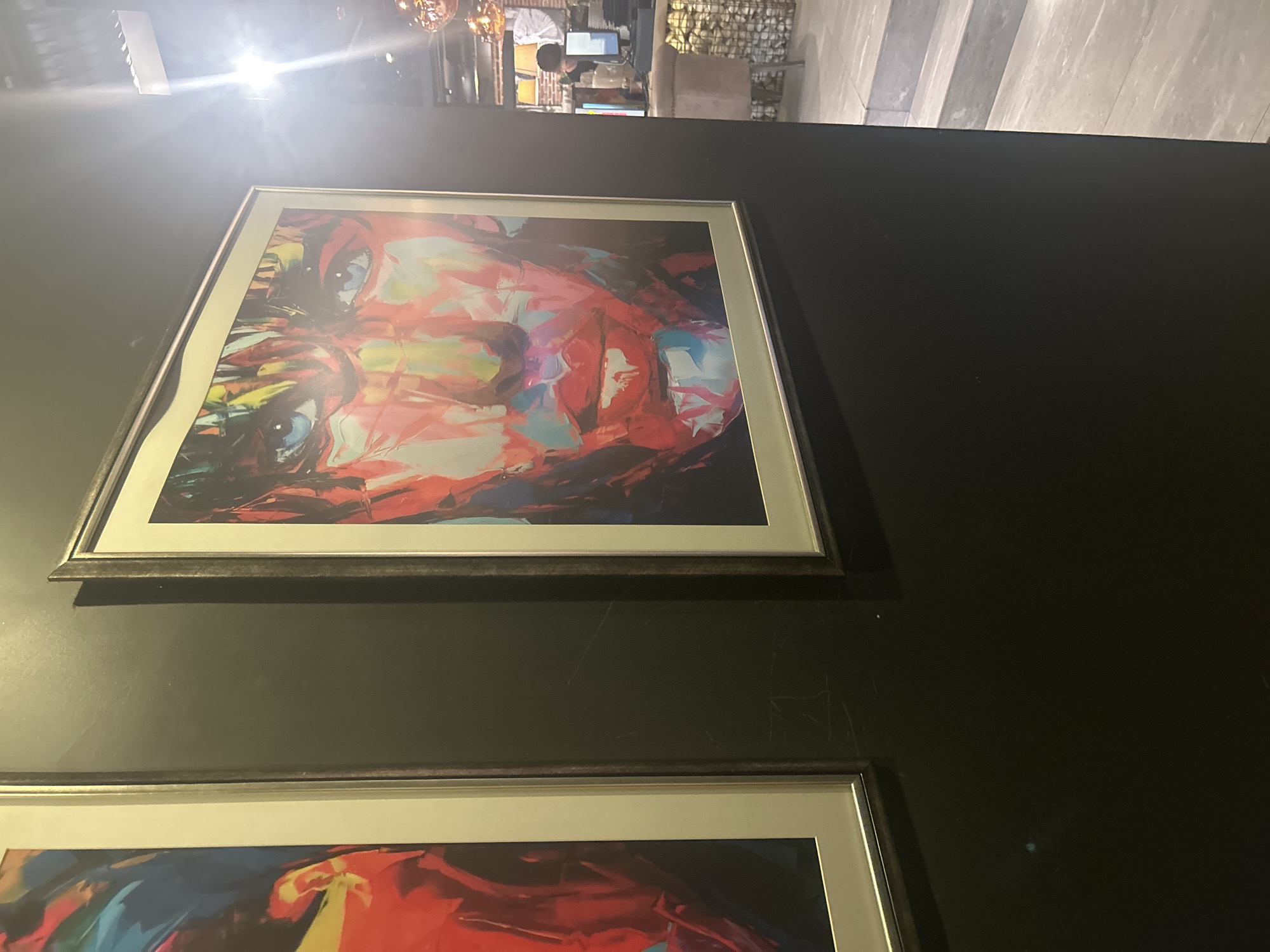 The image shows a close-up of two framed abstract portraits hanging on a dark wall. The portrait on the right, closer to the viewer, depicts a face rendered in vibrant colors, with striking blue eyes and red lips. The portrait on the left is partially visible, also appearing to be an abstract face with bold colors. In the background, beyond the dark wall, a dimly lit interior space is visible, featuring a counter with a computer monitor, a chair, and decorative elements like copper-colored lamps. The floor appears to be tiled with a light stone pattern, and there are stairs leading up to another level. There is no discernible indication of the location in Plovdiv, Bulgaria.