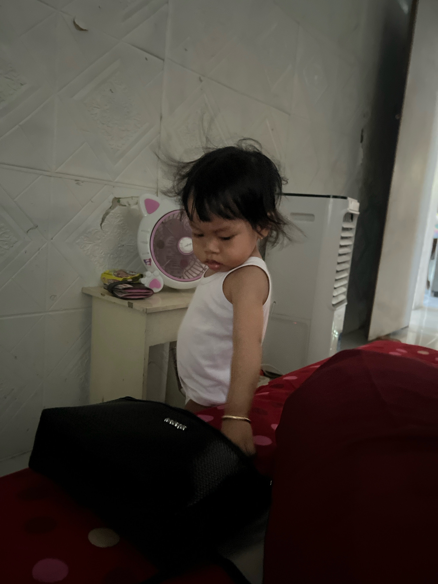 A young child stands near a table with a pink cat-shaped fan and a black purse. The child wears a white tank top and a gold bracelet on their left wrist. The background features a tiled wall and an air conditioner unit. The image is likely taken indoors in a residential setting in Kecamatan Pati, Indonesia.