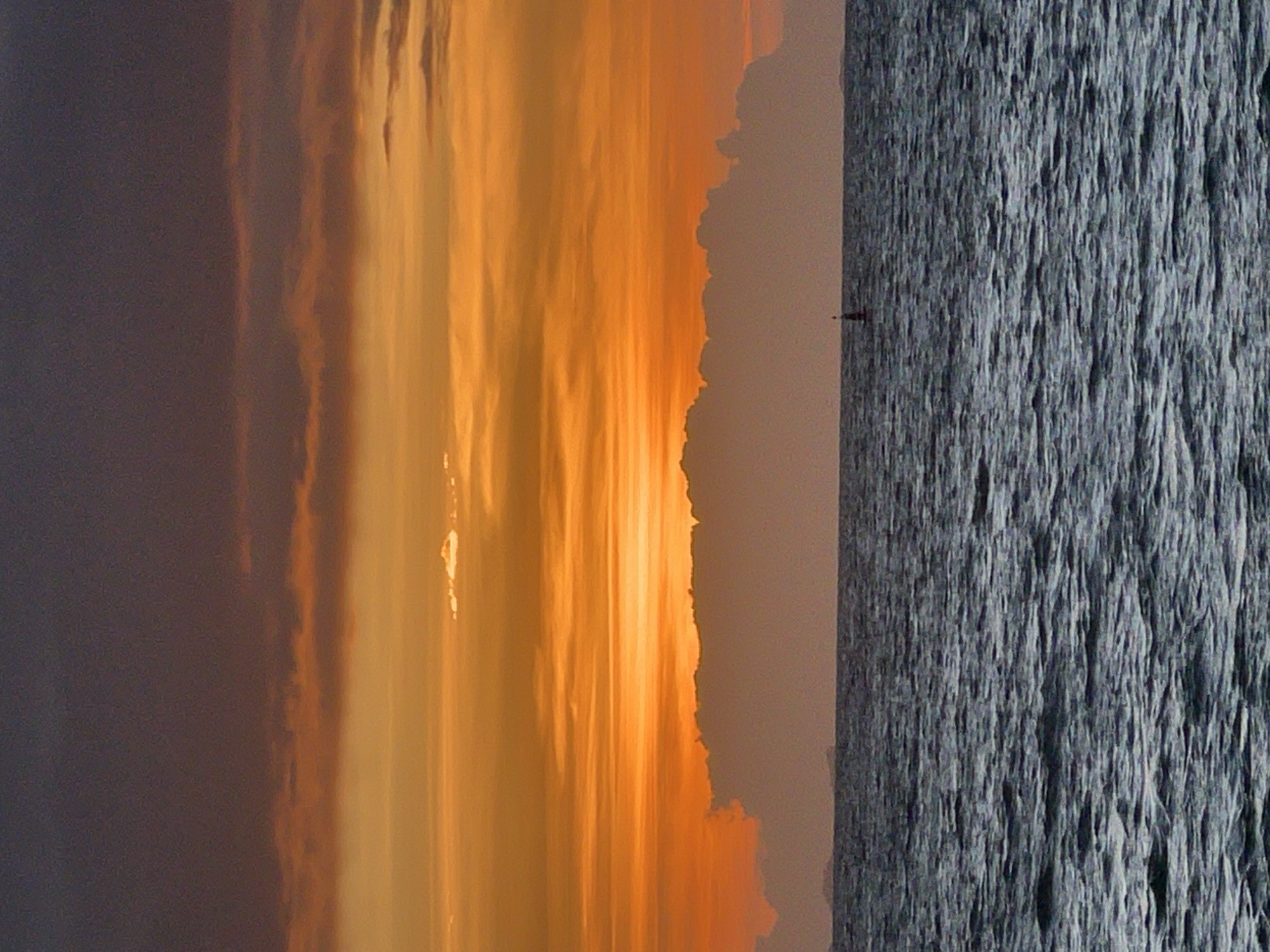 A panoramic view of the ocean at sunset in Ko Pha-ngan District, Thailand, captures the dramatic interplay of light and shadow over the water. The sky is a gradient of deep purples and oranges, transitioning to a vibrant yellow horizon where the sun has just set. Wispy clouds, illuminated by the fading sunlight, stretch across the sky, creating a textured canvas.
Below the celestial display, a dark, choppy sea stretches to the horizon. The water's surface is a tapestry of deep blues and grays, reflecting the subdued light and creating a sense of movement. A few small waves are visible, adding to the dynamic feel of the scene.
In the distance, a faint, dark silhouette of landmasses or islands can be seen on the horizon, barely distinguishable against the twilight sky. Closer to the foreground, a small, red and white buoy stands out against the dark water, marking its presence in the vast expanse. The overall atmosphere is one of tranquility and awe, with the natural beauty of the sunset over the ocean dominating the scene.