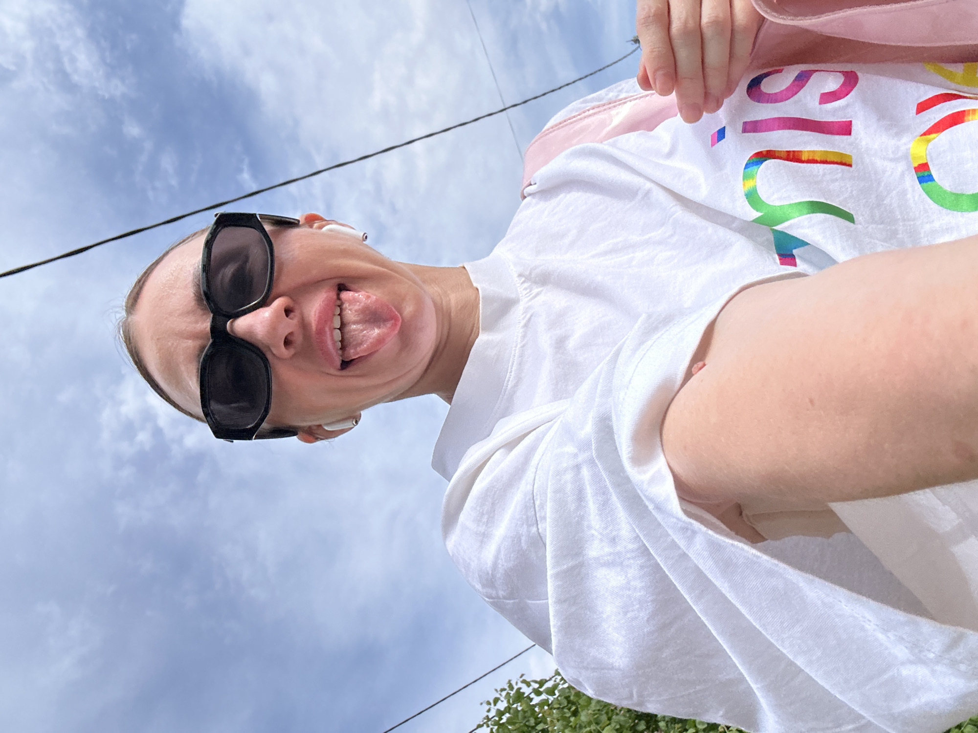 A person with fair skin wearing black sunglasses, white earbuds, and a white t-shirt with colorful lettering is holding a camera or phone and taking a selfie from a low angle. The person is sticking out their tongue. Behind the person is a cloudy sky and electrical wires. Green foliage is visible in the lower corner of the frame. The location cannot be determined from the image.