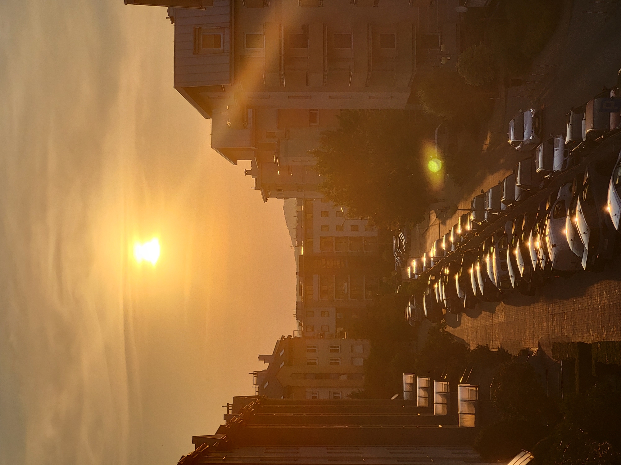 This is a vertical, eye-level, medium shot of a city street at sunset. The sun, a bright white orb with a halo effect, is in the upper left quadrant of the frame, casting a warm, orange glow over the scene. The sky is a hazy orange with streaks of cloud.
On the left side of the frame, a dark apartment building with multiple windows runs vertically. To its right is a street lined with parked cars, most of which are dark-colored. The cars are parked on both sides of a brick-paved street, which curves slightly to the right as it recedes into the distance.
On the right side of the frame, a series of apartment buildings with pitched roofs and visible windows are stacked in the background. A large tree with dark green foliage is situated between the parked cars and the apartment buildings.
The light from the setting sun creates long shadows on the street and reflects off the cars, highlighting their shapes and the street surface. A single white car is driving down the street in the mid-ground. The overall atmosphere is peaceful and serene.