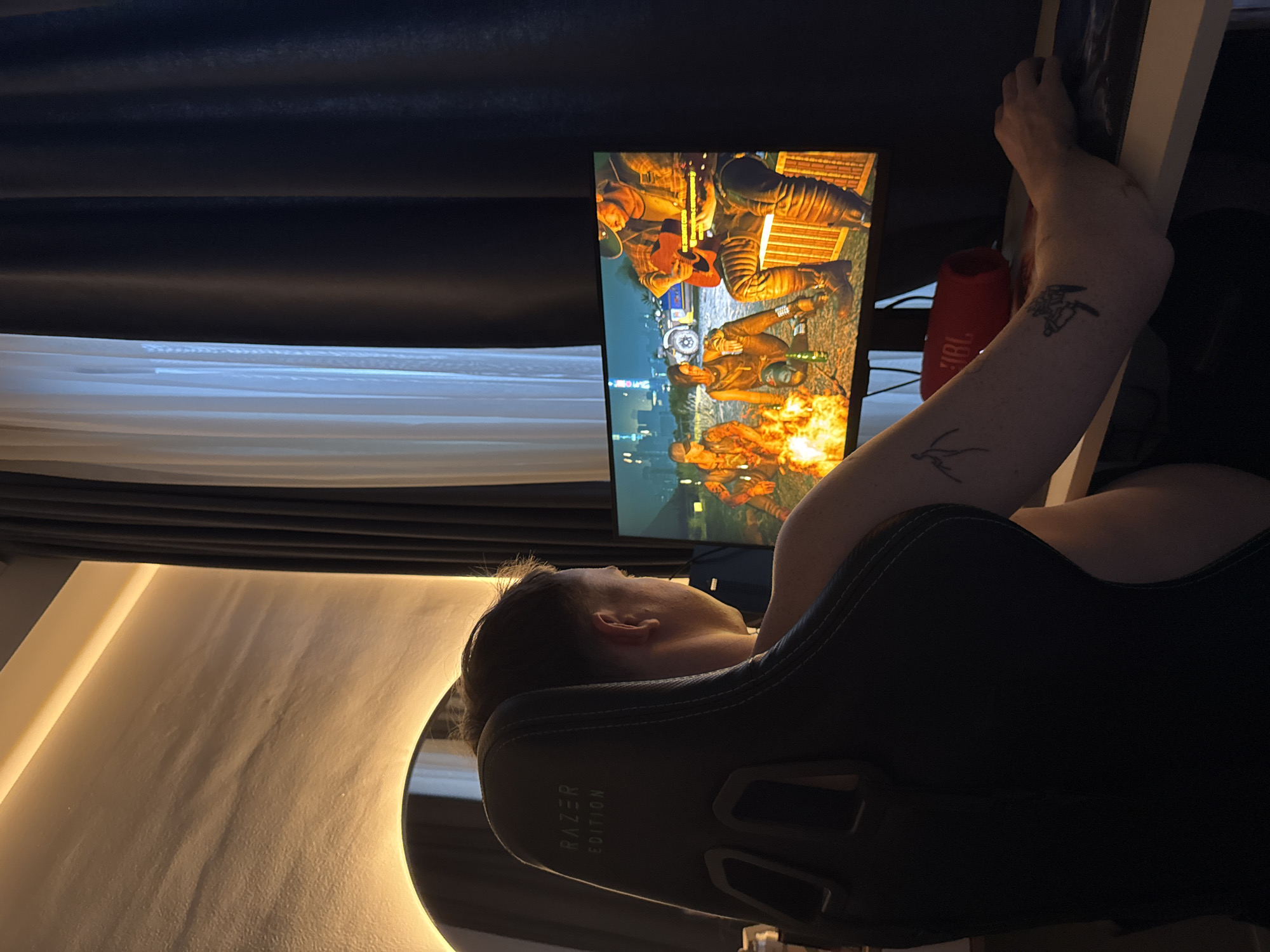 A person is reclining in a black Razer Edition gaming chair, facing a monitor displaying a scene with multiple figures wearing attire suggesting a military or historical theme. The person has fair skin and visible tattoos on their arm. A red JBL speaker is partially visible behind the person's arm. The room is dimly lit with dark curtains and a strip of light on the left side of the frame. It is not possible to determine the city or location from the provided image.