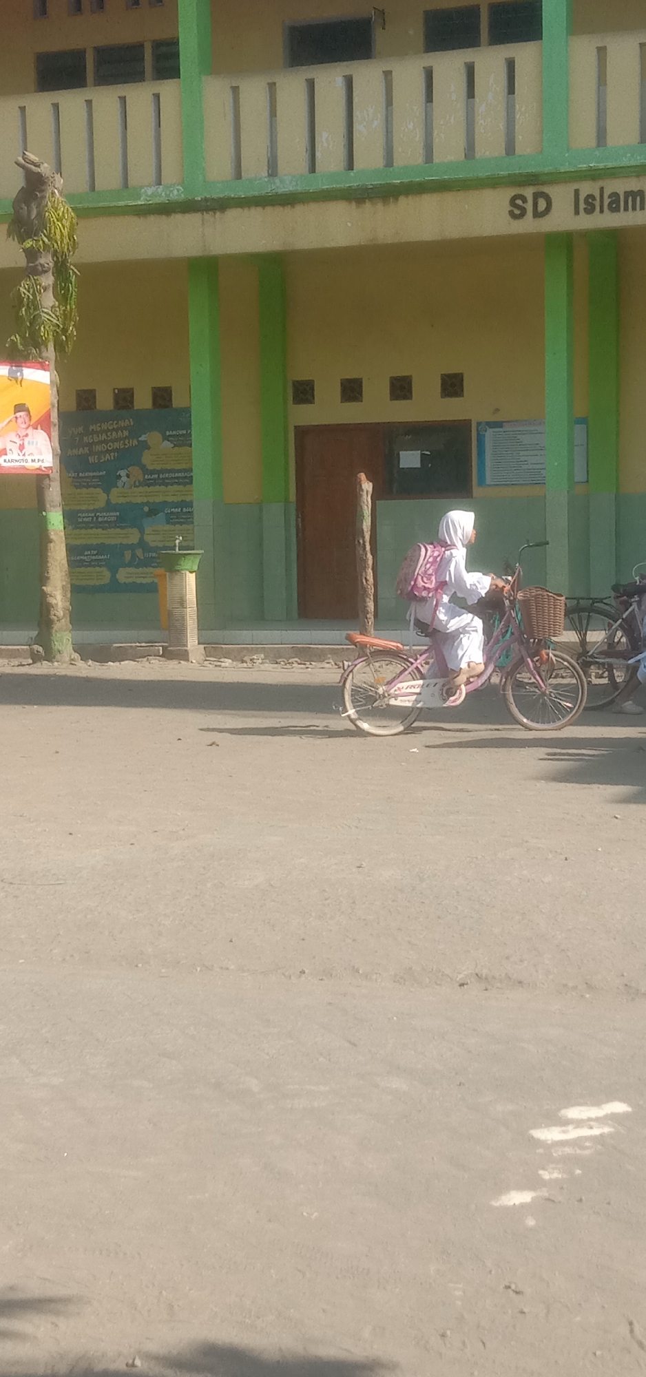 In the city of Sukorejo, Indonesia, a young girl in a white hijab and school uniform rides a pink bicycle down a paved road. She wears a pink and white backpack. The bicycle has a basket on the front, and another bicycle is partially visible behind her. The girl is positioned in the right half of the frame. In the background, a pale yellow building with green trim stands. The building has multiple windows, some of which are covered by louvers. A sign on the building reads "SD Islam." A poster with an image of a man in a uniform and text in Indonesian is attached to a tree on the left side of the image. The ground in front of the building is a sidewalk, and the road surface is asphalt. Sunlight casts shadows on the ground.