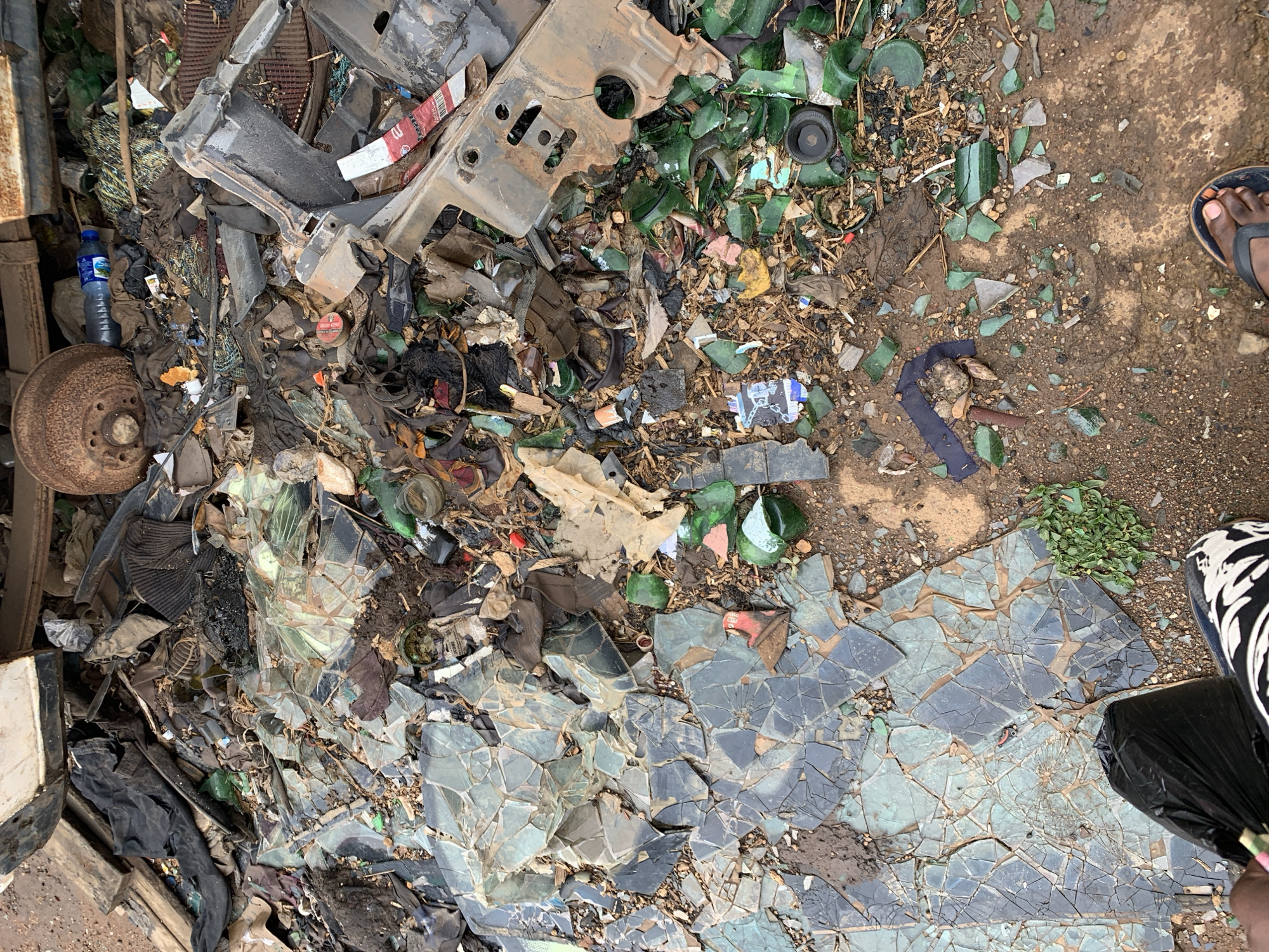 The image shows a littered area in Rafin Par, Nigeria. It is covered with various types of waste including metal scraps, broken glass (mostly green), plastic bottles, general trash, and what appears to be pieces of broken tile or similar material.
On the upper right of the image there is a patch of brown dirt, interspersed with small pieces of glass. The feet of two individuals are visible; one wearing sandals and the other sneakers. The scene suggests an environment marred by indiscriminate dumping of waste.
