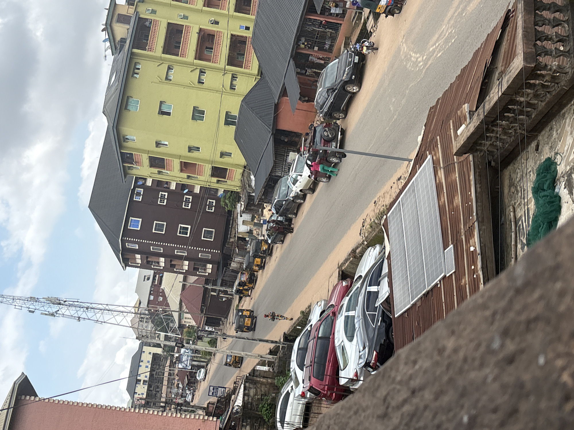 The image depicts a street scene in Nkpor, Nigeria, with a focus on several parked cars and buildings. In the foreground, a rusty corrugated iron roof with solar panels is visible, with a concrete railing and a green net. The street itself is paved and shows signs of wear. Several cars are parked along the side of the road, including a white sedan, a red SUV, a silver sedan, and a black sedan. Further down the street, a person in a camouflaged uniform is walking. A cell phone tower stands tall in the background, and multiple multi-story buildings line the street, some with brightly colored facades, like the prominent lime green building. Several tricycles, commonly used for public transport in Nigeria, are also present on the road. The sky is partly cloudy.