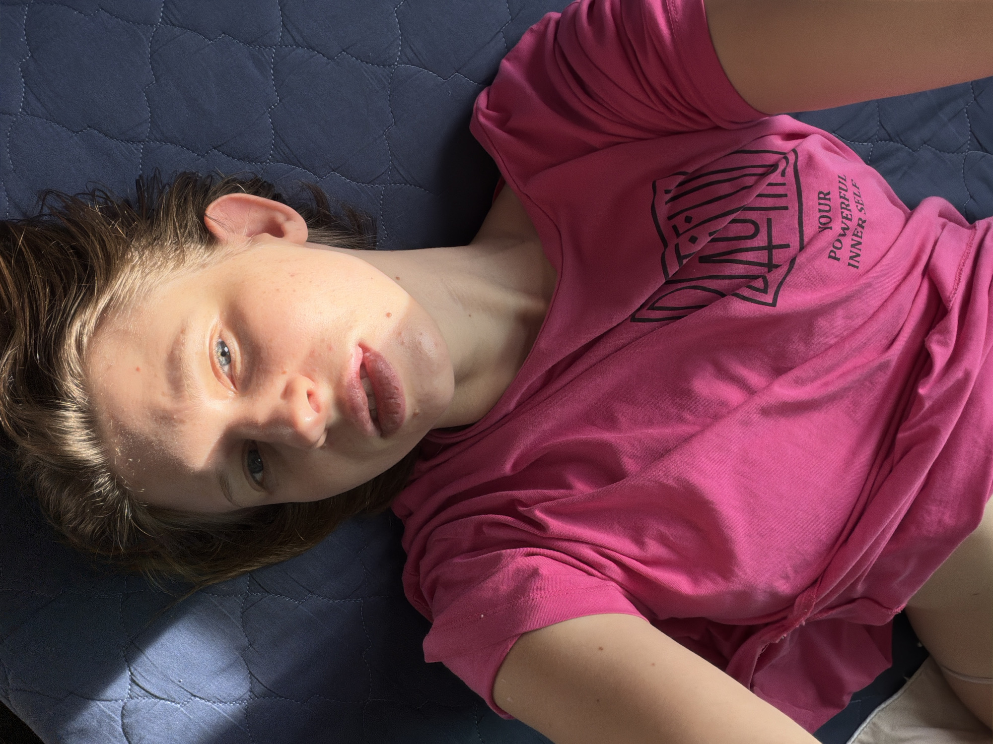 A woman lies on a blue quilted surface, her head turned towards the camera. She wears a bright pink t-shirt with a graphic design and text that reads "YOUR POWERFUL INNER SELF". Sunlight streams across her face, highlighting sweat on her forehead and casting shadows. Her eyes are open and looking upwards, and her mouth is slightly parted. The background is a dark blue quilted fabric. The scene appears to be indoors, possibly in a bedroom, in Dongducheon-si, South Korea.