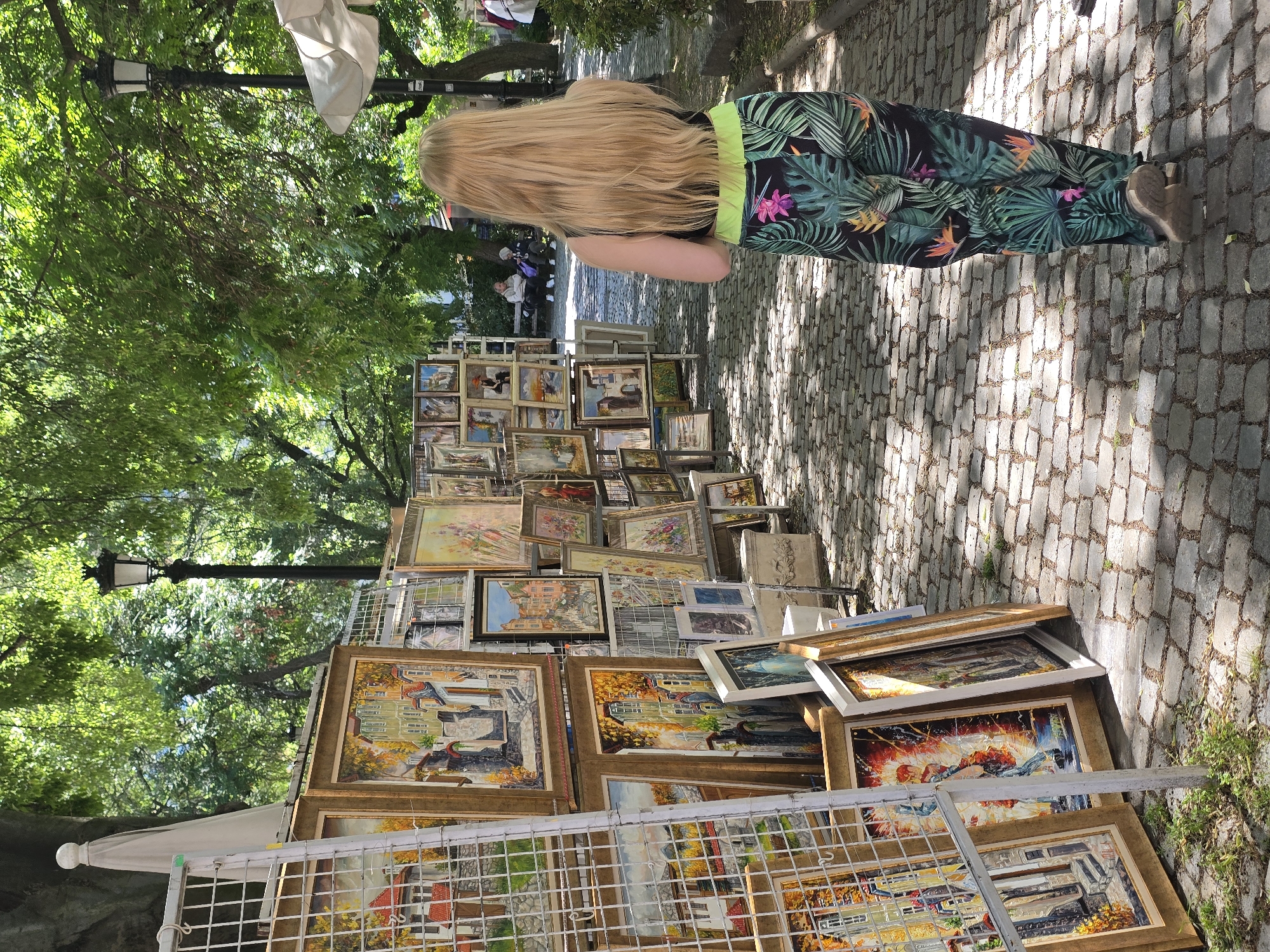 A street scene in Plovdiv, Bulgaria, features an outdoor art exhibition displayed along a cobblestone street. A fair-skinned woman with long blonde hair stands viewed from the back, wearing floral print pants with a lime green waistband and grey shoes. She appears to be contemplating the art. The art display includes numerous framed paintings leaned against a wire grid display rack. The artwork covers landscapes and cityscapes. Trees and lampposts are visible along the street in the background. The lighting suggests a sunny day.