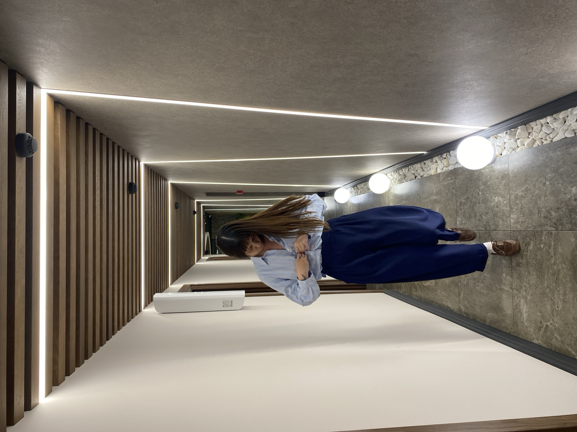 A full-body, medium-shot image shows a woman with long, dark hair and bangs walking down a hallway, with her hands occupied with the buttons of her light blue striped button-down shirt. She is wearing dark blue wide-leg pants and brown loafers with white socks. The hallway features a slatted wooden ceiling with integrated LED strip lighting, and textured beige walls with vertical LED strip lighting. The floor is a dark gray marbled tile. Several spherical white lights are placed along the right side of the hallway, interspersed with white stones. The scene is indoors, in what appears to be a modern building. The location is not identifiable as Votkinsk, Russia.