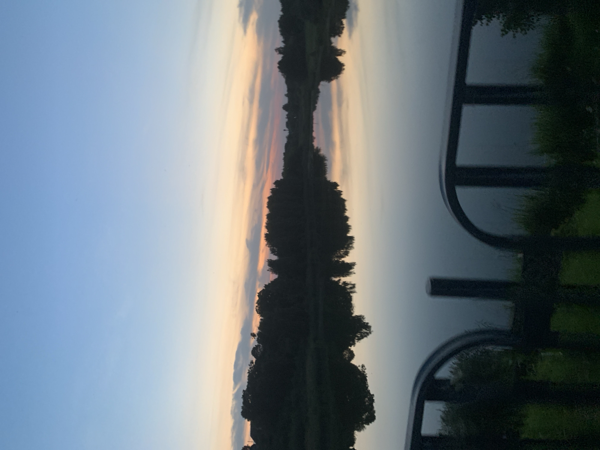 A wide shot captured from a low angle shows a serene body of water reflecting a dusky sky. The sky transitions from a pale blue at the top to soft orange and pink hues near the horizon, suggesting either sunrise or sunset. Silhouetted trees line the far bank of the water, creating a dark, undulating border against the vibrant sky. In the foreground, a dark, blurred railing, possibly part of a bridge or walkway, frames the lower portion of the image. The water itself is calm and glassy, mirroring the sky and the distant treeline with remarkable clarity. The overall impression is one of tranquility and natural beauty. The image is not specific to any particular location within the city of Kapyĺ, Belarus, but represents a generic natural landscape that could be found in or near the city.