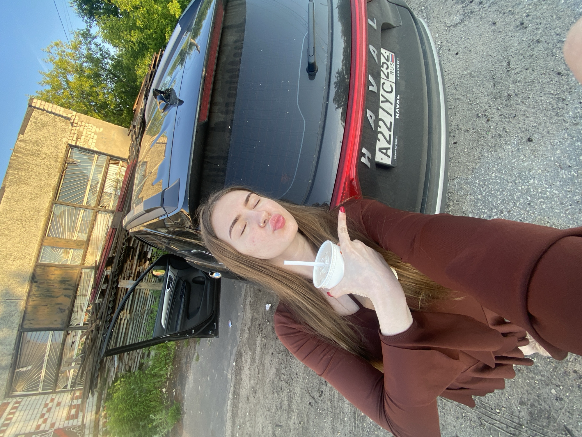 A young woman with long brown hair and closed eyes is posing with a cup and straw in her right hand, puckering her lips and pointing towards the rear of a black SUV parked outdoors. She is wearing a dark red, ruffled long-sleeved shirt. The car's rear hatch is open, and the license plate reads "A227YC 254 RUS", indicating it is registered in Nizhny Novgorod, Russia. The background shows a brick building with boarded-up windows, stacked wooden planks, and green foliage under a clear blue sky. The ground is paved with asphalt or concrete. The overall scene appears to be in a semi-industrial or commercial area.