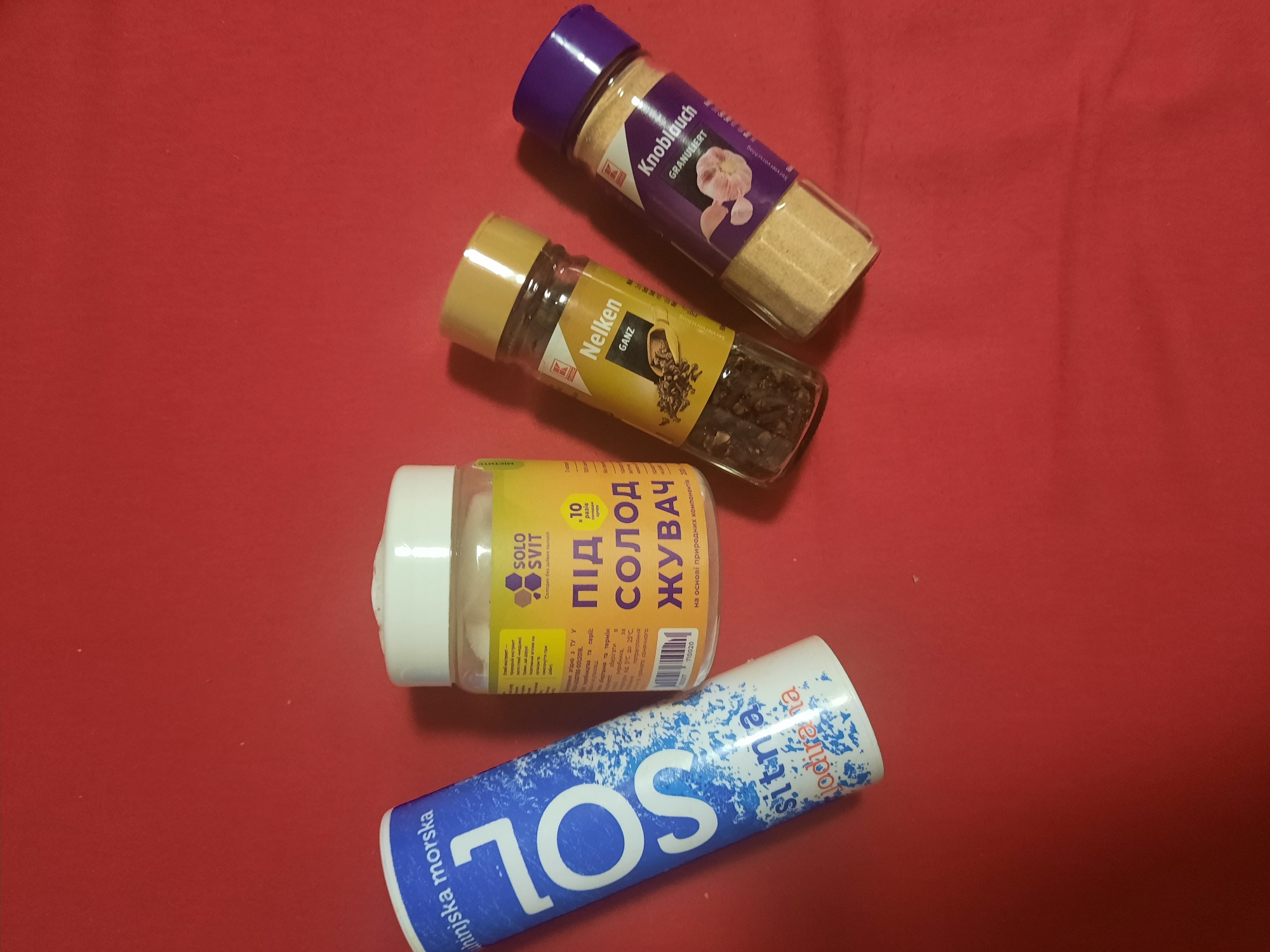 The image displays a collection of four food-related items arranged on a red, textured surface, likely fabric. The items are positioned in a loose, diagonal line from the bottom left to the top right of the frame.
On the far left is a tall, cylindrical container of "SOL" brand iodized salt. The container is predominantly white with a blue and white speckled design and large blue lettering that reads "SOL". Text in Ukrainian and Cyrillic script is visible, including "sitna" and "jodirana", indicating it is iodized salt.
To the right of the salt container is a clear, round plastic jar with a white lid. This jar contains a white granular substance and has a yellow label with Cyrillic text. The prominent text on the label reads "ПІД СОЛОД ЖУВАЧ" (Pod Solod Zhuvach), which translates to "For Sweet Chewing". It also features the brand name "SOLO SVIT" and mentions it is made from natural components.
Next to the jar is a clear glass spice grinder with a yellow plastic cap. The label on this grinder is in German and reads "Nelken GANZ", indicating whole cloves. The grinder contains whole dark cloves.
On the far right is another clear glass spice grinder, this one with a purple plastic cap. The label on this grinder is also in German and reads "Knoblauch GRANULIERT", indicating granulated garlic. The grinder contains a light tan, granular powder.
The scene is devoid of any people or animals. The background is a solid, vibrant red. The lighting appears to be overhead, casting subtle shadows. The image does not provide any information about its location in Rosenheim, Germany.