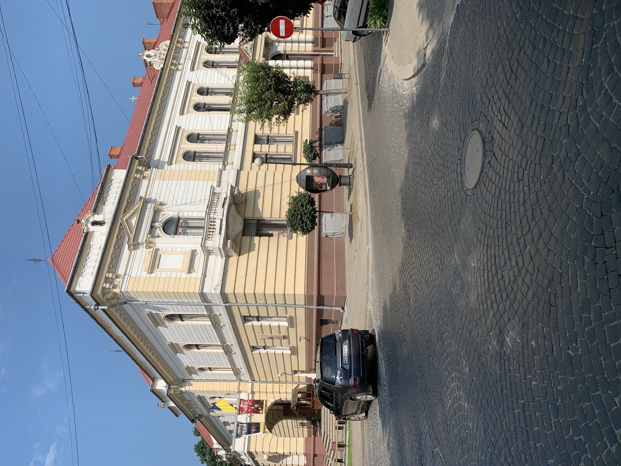 The image shows a street scene in Kolomyia, Ukraine, on a sunny day. A dark-colored SUV is parked on a cobblestone street in front of a building with a yellow and white facade and a red tiled roof. The building appears to be a public institution, possibly a theater or cultural center, as indicated by banners displayed near the entrance. There are trees and a street lamp on the sidewalk. A red and white "no entry" sign is visible on the right side of the street. The sky is clear and blue.