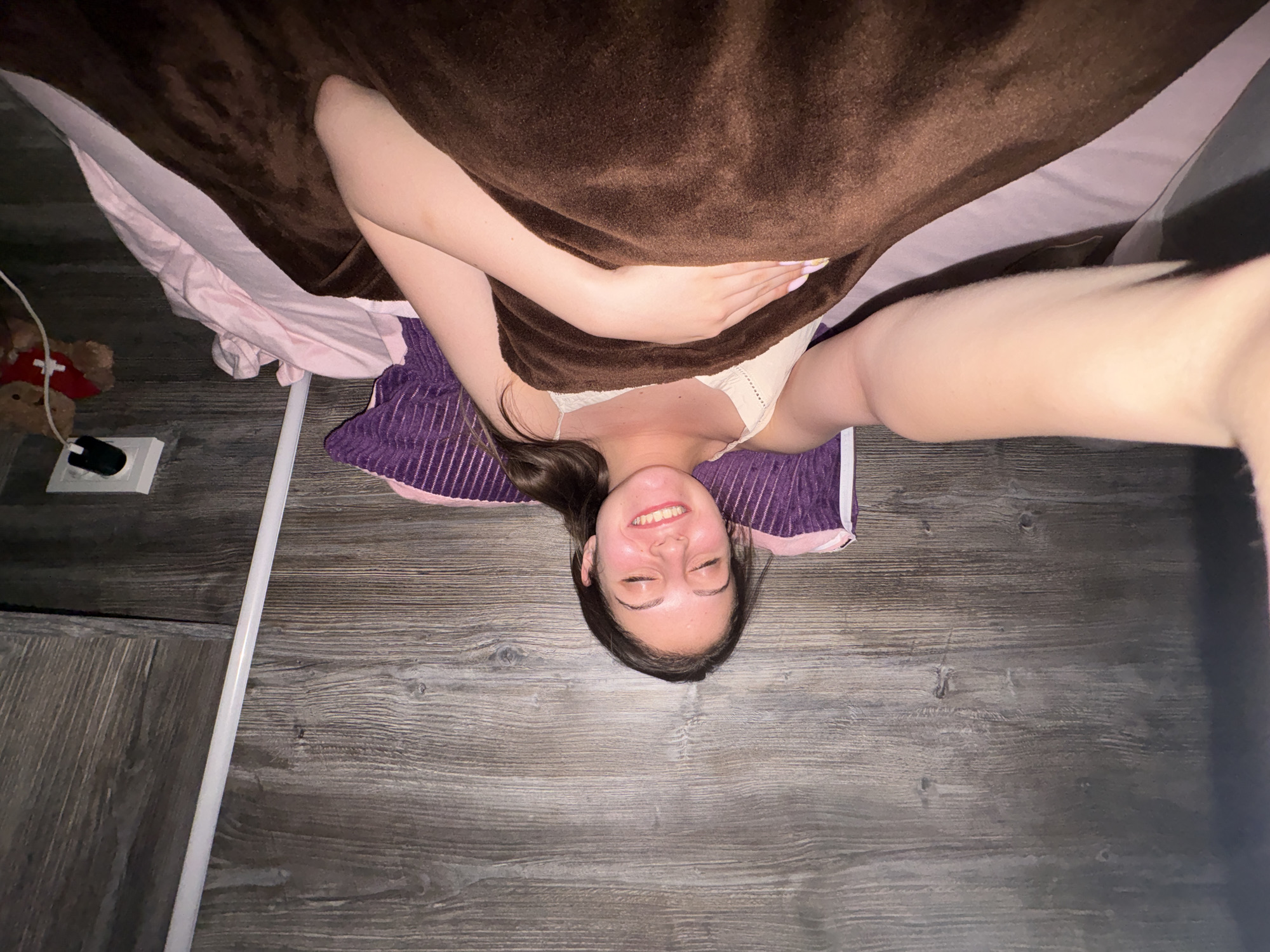 A young woman is shown from an overhead perspective, lying on her back with her head towards the bottom of the frame. She has long, dark hair and is smiling broadly with her eyes closed, conveying happiness or contentment. She is wearing a light-colored bra or top. A large, brown, fuzzy blanket or towel partially covers her body, with one arm bent and her hand resting on her stomach area, and the other arm extended towards the camera. A purple striped cushion or pillow supports her head, with light pink fabric visible underneath the brown blanket.
The setting appears to be an indoor room with a dark wooden floor. In the bottom left corner, a small brown teddy bear wearing a red accessory is visible next to a white power outlet and a black computer mouse with a white cable. A long white linear object runs diagonally across the floor. There is no visible text in the image. This scene takes place in Bulgarovo, Bulgaria.