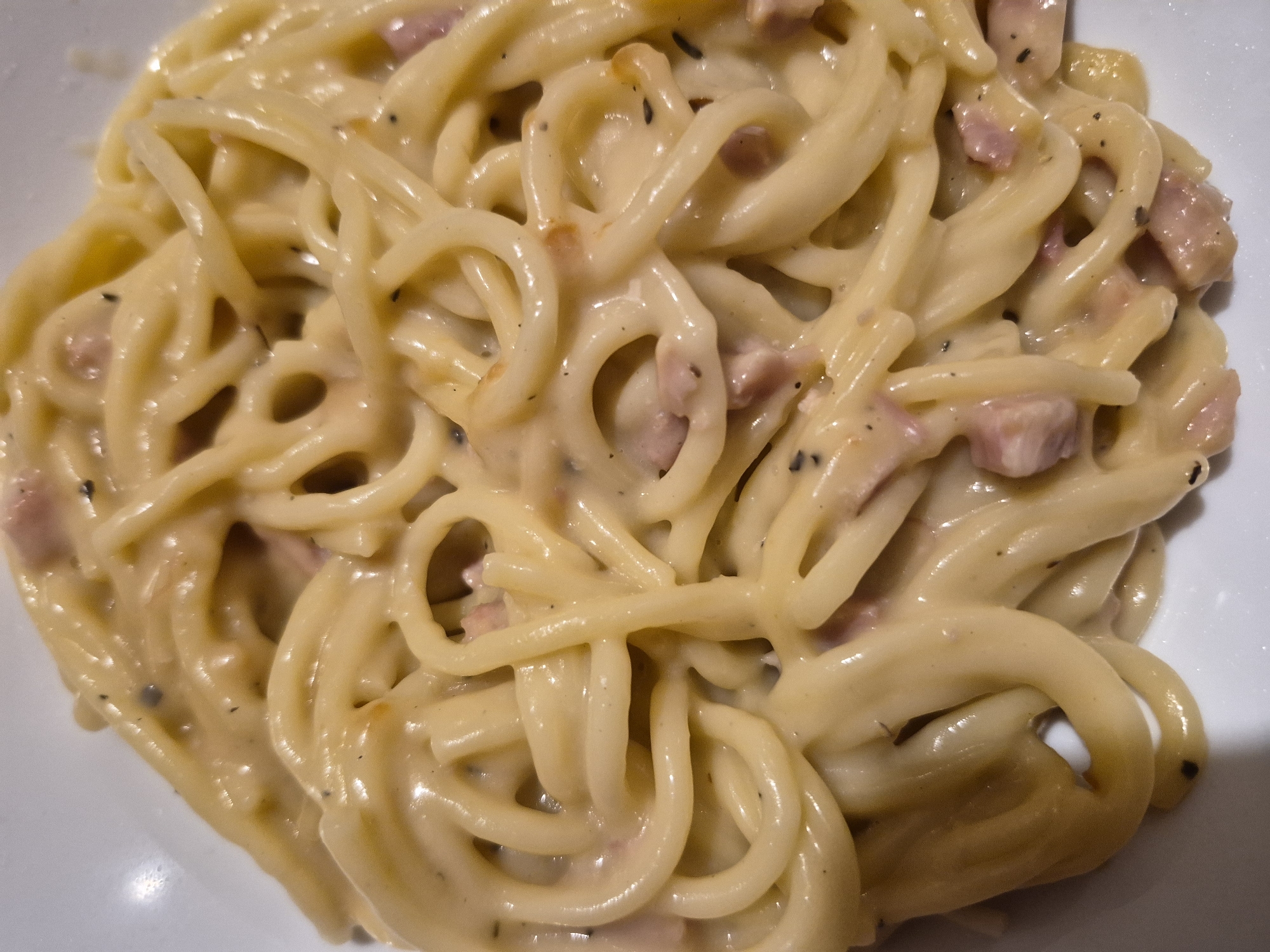 A close-up, top-down shot shows a dish of spaghetti carbonara. The pasta is coated in a creamy sauce with visible pieces of ham and black pepper. The dish is served in a white bowl, with a portion of the bowl visible in the lower left corner. The background is blurred and out of focus. There are no people or animals in the image. The scene is likely a restaurant or home kitchen in Sandwell, United Kingdom.