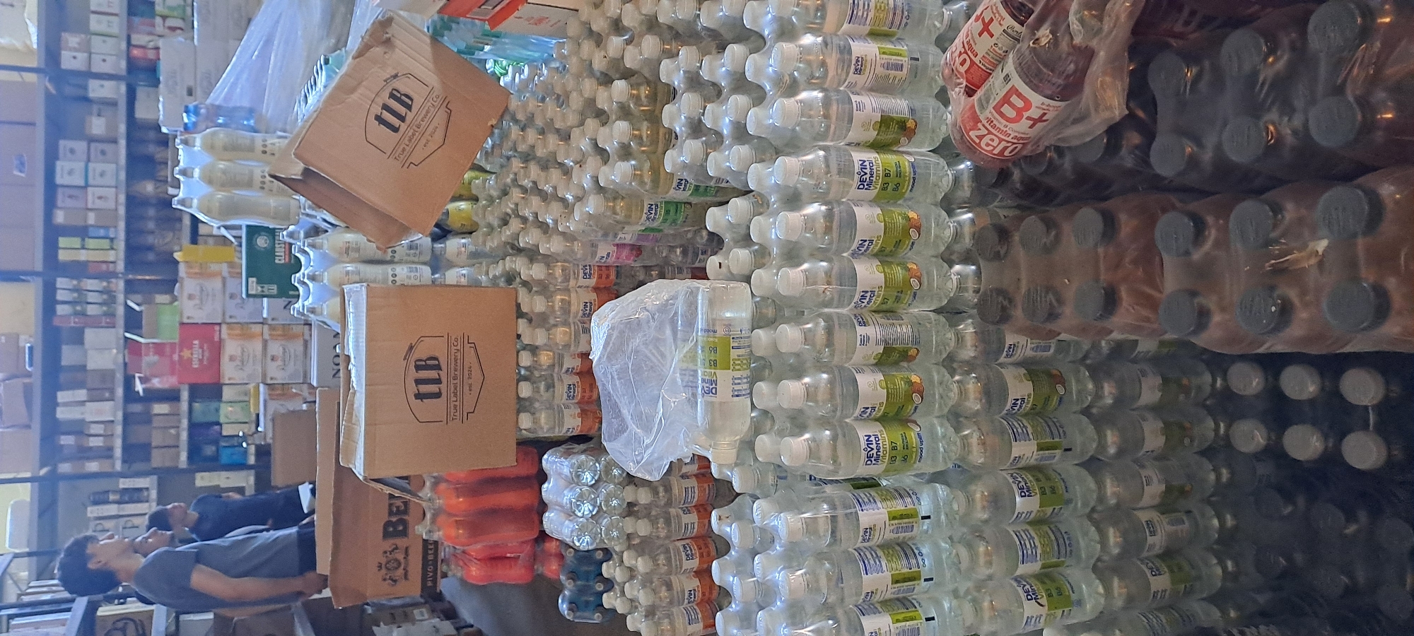 The image depicts a storage room filled with bottled beverages, likely a warehouse or distribution center. The room is densely packed with cases and stacks of bottles of various sizes and types. In the foreground, numerous clear plastic bottles of water and other beverages with white and green labels are stacked on shelves and on the floor. Some bottles have labels indicating "DEVIN Mineral Vitamins." To the left, there are stacks of red-colored bottles and clear bottles. In the center of the image, two cardboard boxes with the "TLB True Label Brewery Co." logo are visible. In the background, shelves are stocked with more boxes and bottles, some appearing to be beer crates. Two young men are present in the left side of the image, standing and looking towards the shelves. The overall environment appears to be utilitarian and organized for storage. The presence of "Velika" in the prompt suggests this scene is located in Velika, Bulgaria.
