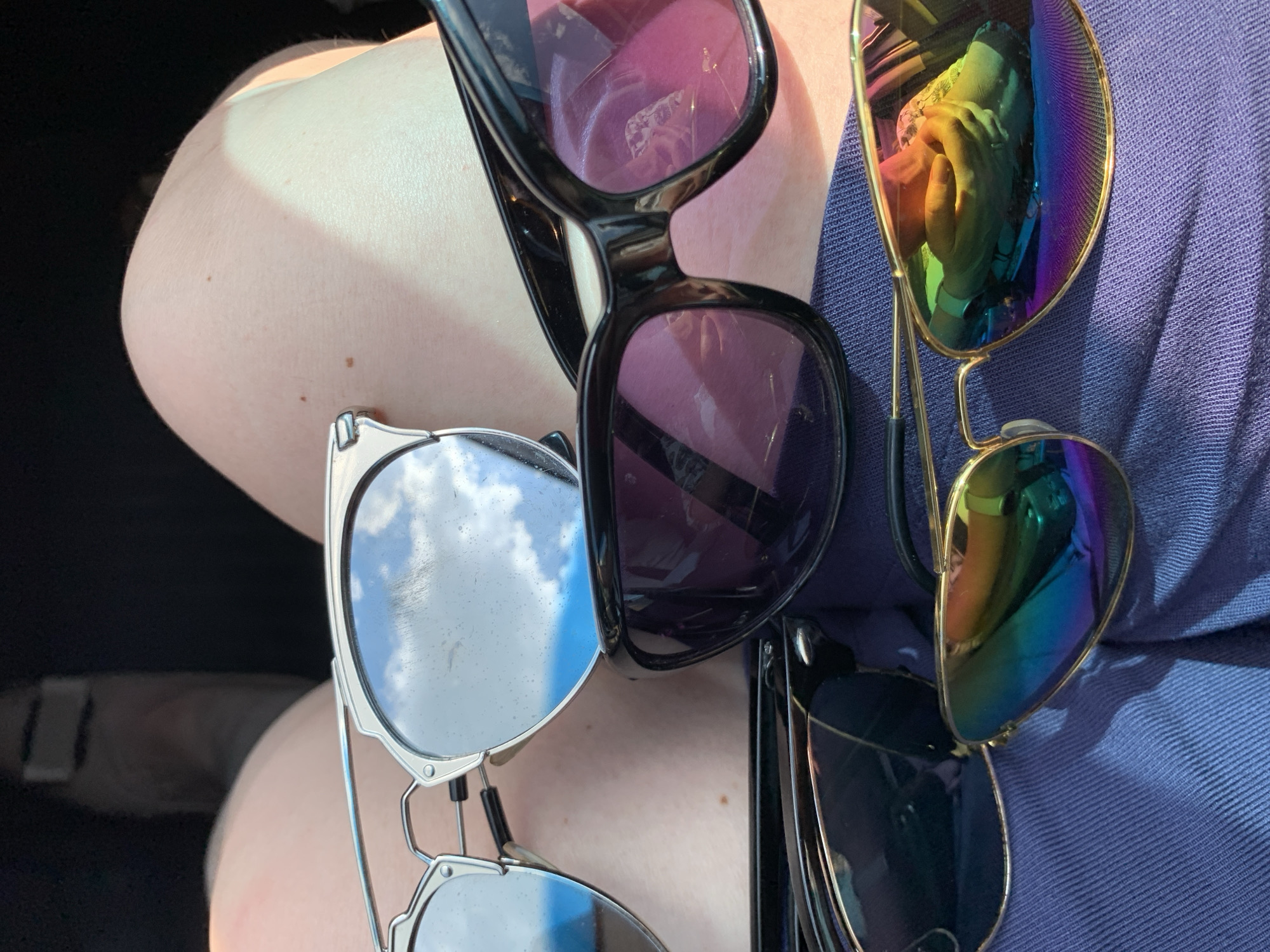 The image shows a close-up, eye-level shot of four pairs of sunglasses resting on blue fabric, likely clothing. The sunglasses are of various styles and colors. The pair closest to the viewer has a gold frame and mirrored lenses reflecting a rainbow of colors. Another pair has a black frame and dark lenses with a pinkish tint. A third pair has a silver frame with mirrored lenses that reflect a blue sky with white clouds. The fourth pair, partially obscured, appears to have a dark frame and dark lenses. The background is out of focus but suggests an indoor setting, possibly a car interior, with hints of a person's legs visible. There is no indication of the location being in Азерскі сельскі Савет, Belarus.