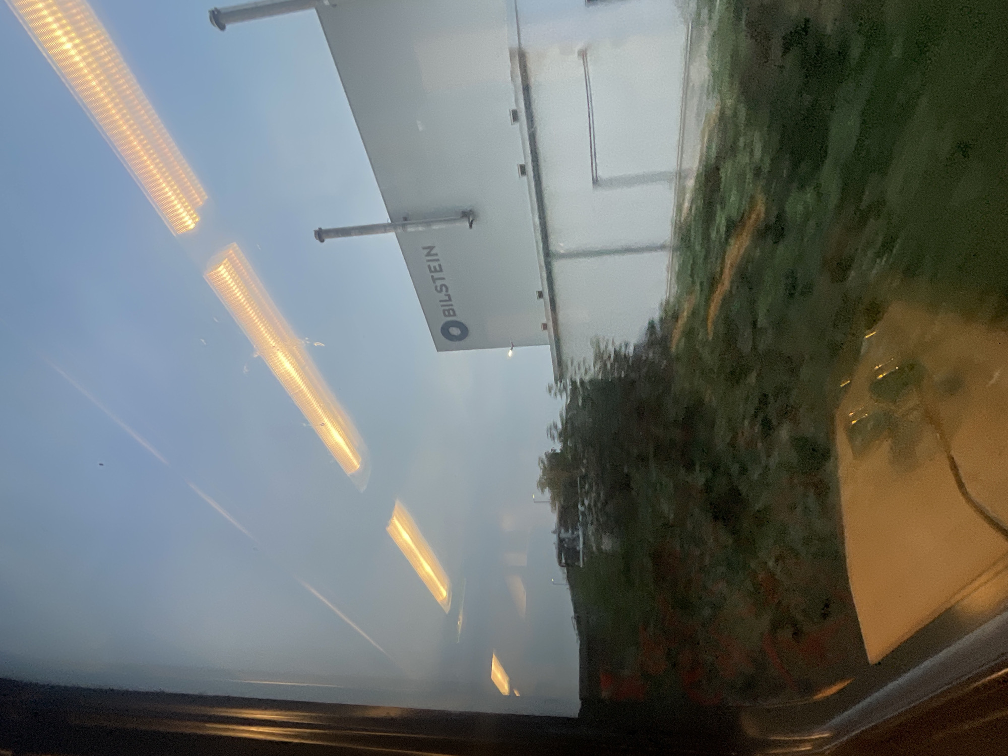 The image, taken from inside a vehicle, shows a view through a window. In the distance, a large, white industrial building with the "BILSTEIN" logo is visible. The building has several smokestacks. The sky is a clear, pale blue, with several overhead lights reflecting on the window. In the foreground, green foliage and a patch of orange-brown dirt are visible on a slope. The interior of the vehicle, including a light-colored surface and part of a dark frame, is also reflected in the window. The scene appears to be in an industrial area. The Bilstein Group has a facility located in the city of Hagen, Germany.