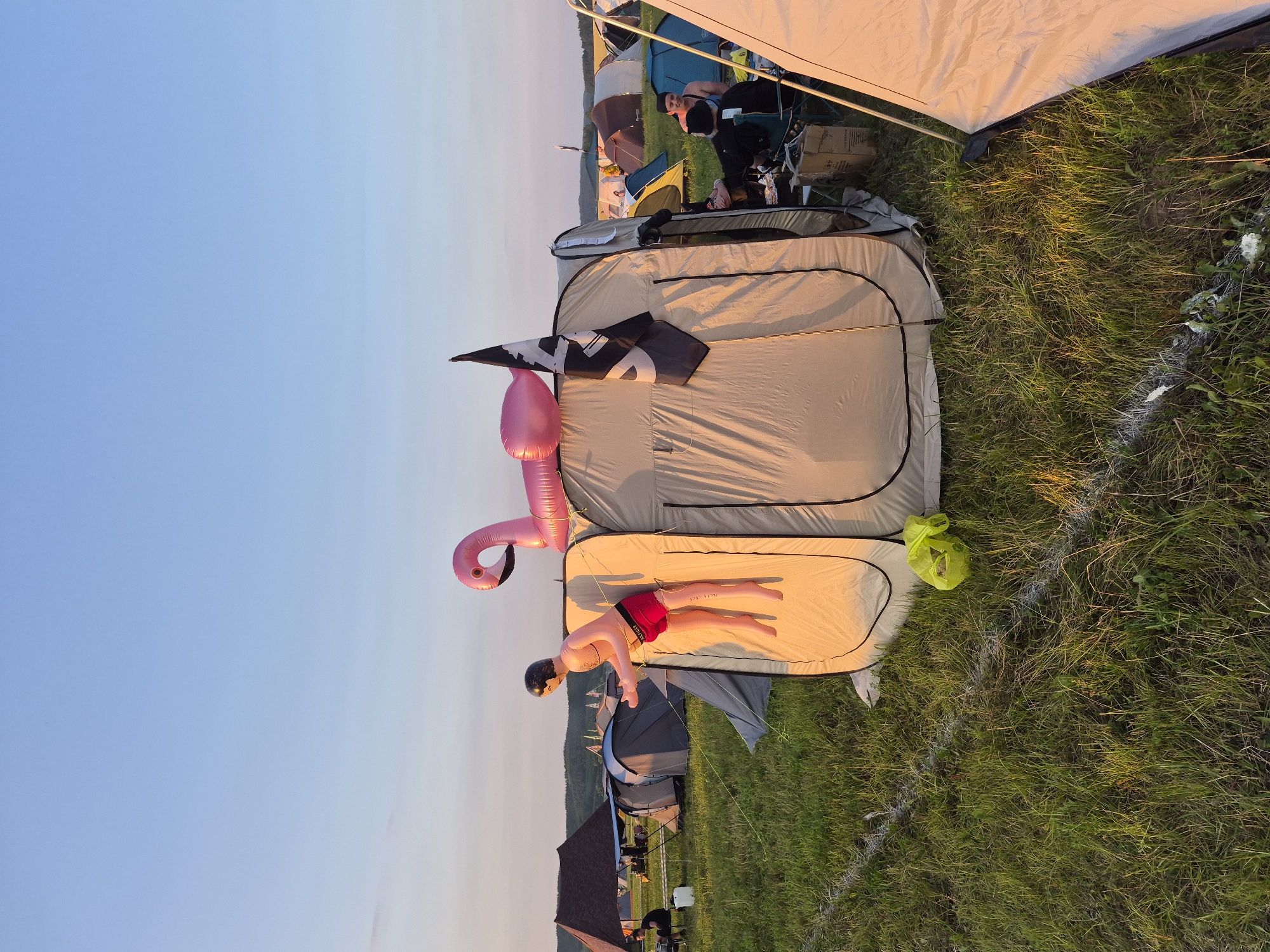 In Lagunovo, Russia, a campsite is set up on a grassy field under a clear sky at sunset. A large, tan pop-up tent is the focal point, with an inflatable pink flamingo resting on its roof. A mannequin, dressed in red swim trunks and with an artificial tan, is propped up against the side of the tent, appearing to emerge from it. A black and white flag with a skull symbol flutters from the tent's peak. Several other tents and camping structures are visible in the background, along with a group of people sitting near a tent in the middle ground. The ground is marked with a white line, possibly indicating a pathway or boundary. A bright yellow bag lies on the grass near the pop-up tent.