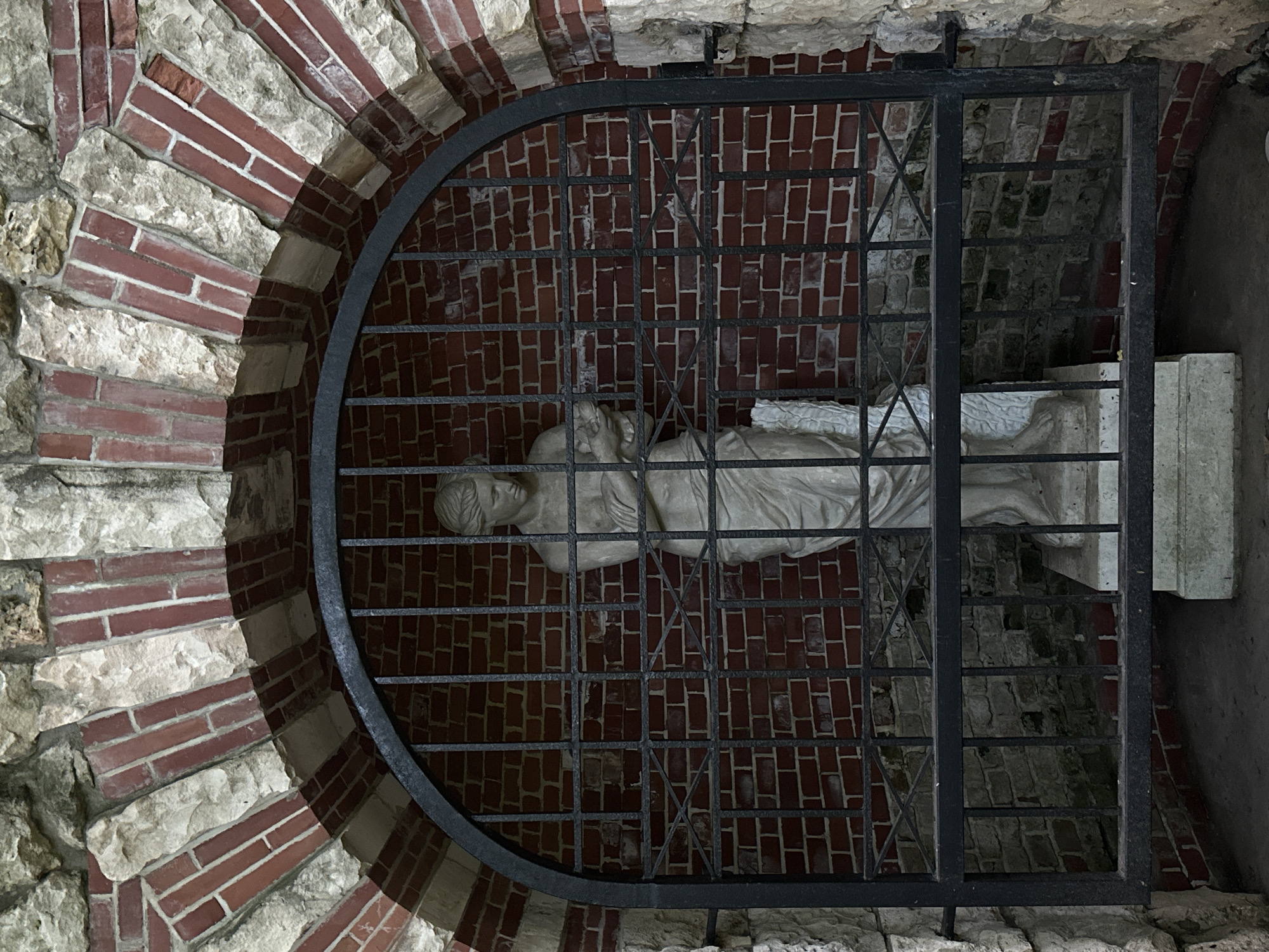 The image shows a white marble statue of a woman holding an infant, enclosed within a dark metal grate. The grate is set within a semicircular arch constructed of red brick and light-colored stone. The statue stands on a pedestal, and behind it, the wall is made of red brick. The overall environment appears to be an outdoor alcove or niche, possibly part of a garden or park. The statue's pose suggests tenderness or mourning, and the grate implies protection or confinement. The scene is dimly lit, contributing to a somber or contemplative atmosphere. This structure is located in Tsaritsyno Park, Moscow, Russia.
