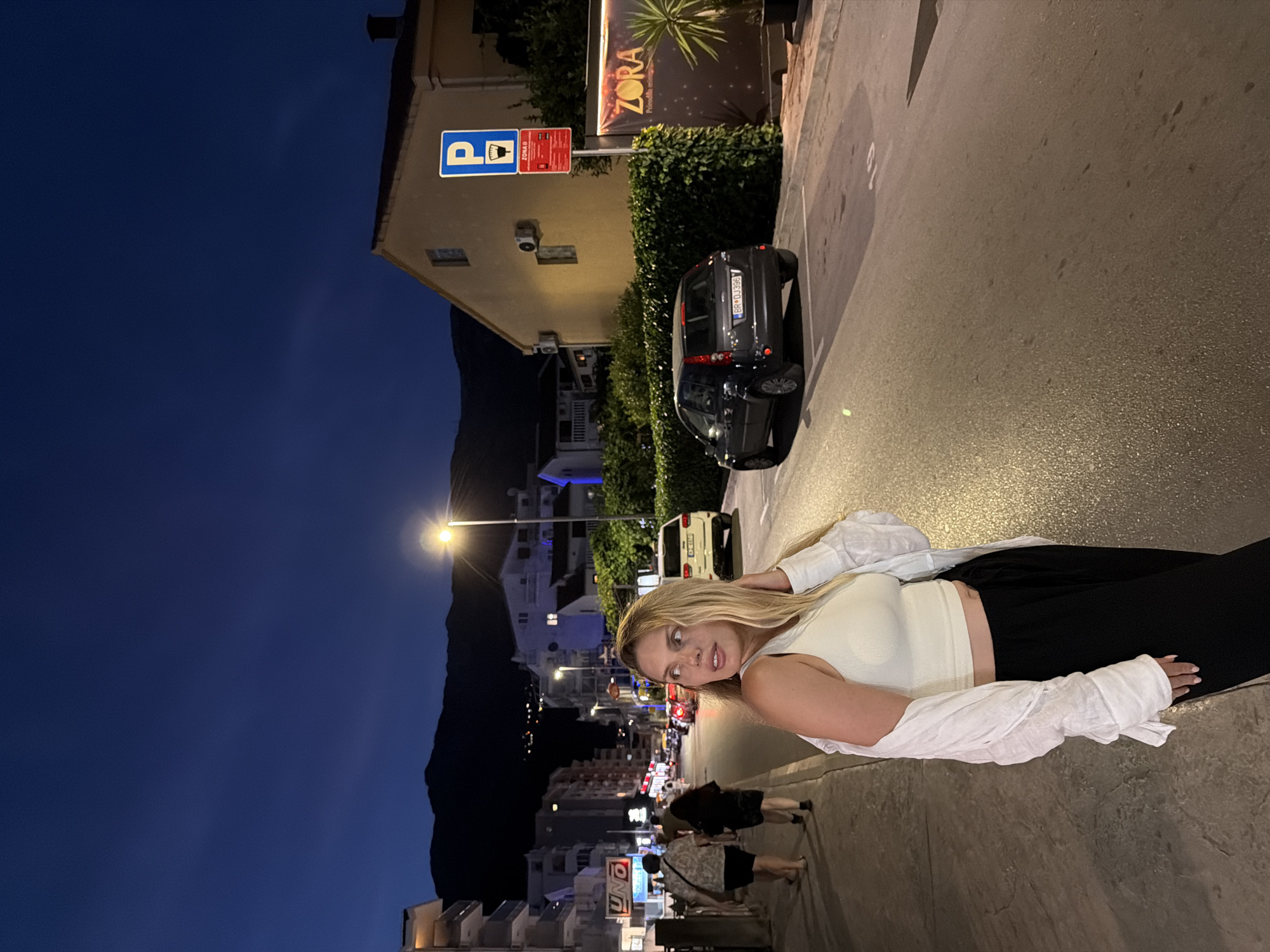 A young woman with blonde hair is standing on a street at night in Budva, Montenegro, looking to her right. She is wearing a white crop top, black pants, and an unbuttoned white shirt draped over her shoulders. The street is paved and has streetlights casting a warm glow. There are buildings lining the street, some with illuminated windows. Several cars are parked along the side of the road, and a few people are walking in the background. A parking sign and a banner advertising "ZORA" are visible on the right side of the image. In the distance, mountains are silhouetted against the dark blue night sky. The scene appears to be a typical street in a town or city at night, with elements of urban infrastructure and leisure.