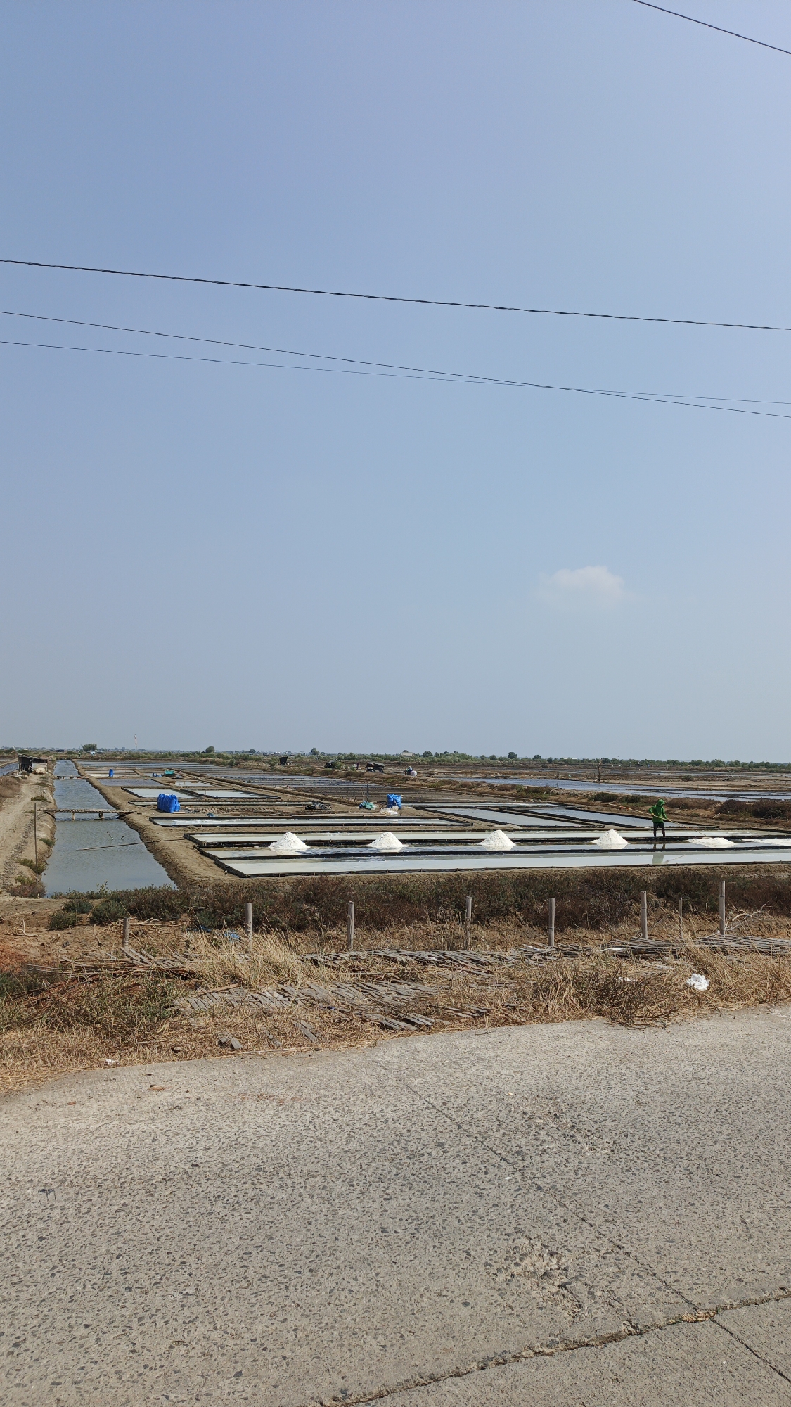 The image depicts a salt field in Krimun, Indonesia, under a clear blue sky. The salt field is divided into numerous rectangular ponds, some of which contain water. Several large piles of harvested salt are visible in some of the ponds. A person in a green shirt is seen raking salt in one of the ponds. In the background, there are scattered structures and vegetation. Two power lines stretch across the upper portion of the sky. In the foreground, a paved road runs along the edge of the salt field, with dry grass and wooden structures lining the embankment.