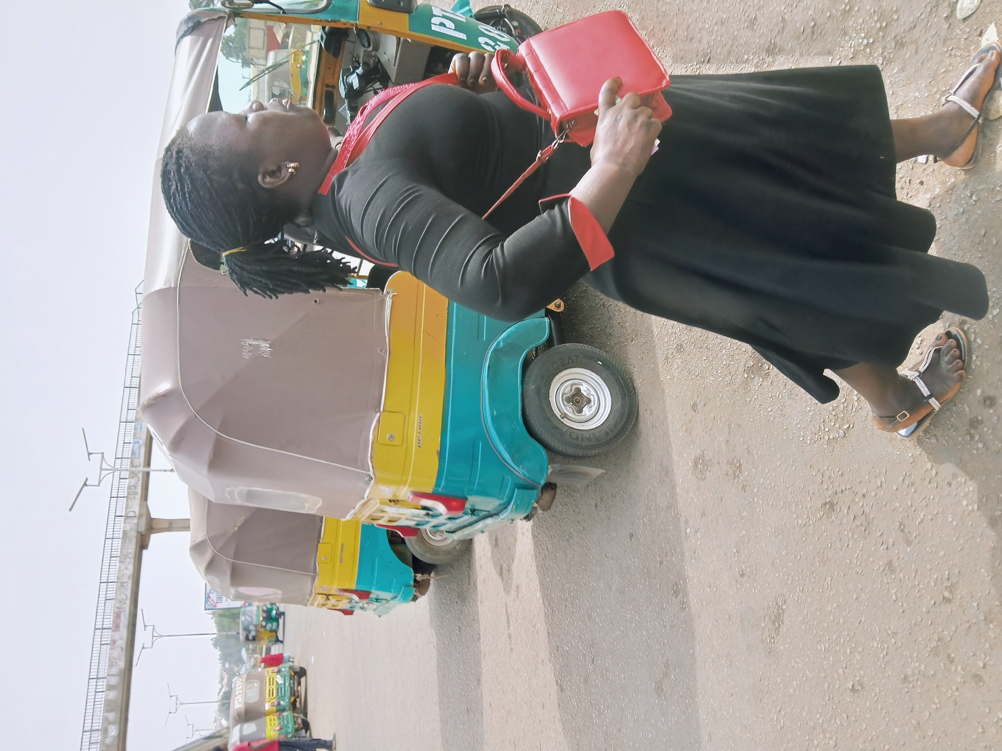 A woman in a long black dress with red trim and sandals stands next to a brightly colored tricycle taxi. She holds a small red purse. The taxis are lined up on a street, likely awaiting passengers. The location is indicated as Jos, Nigeria. In the background, there are other tricycles and a pedestrian overpass. The environment appears to be a busy street in an urban setting.