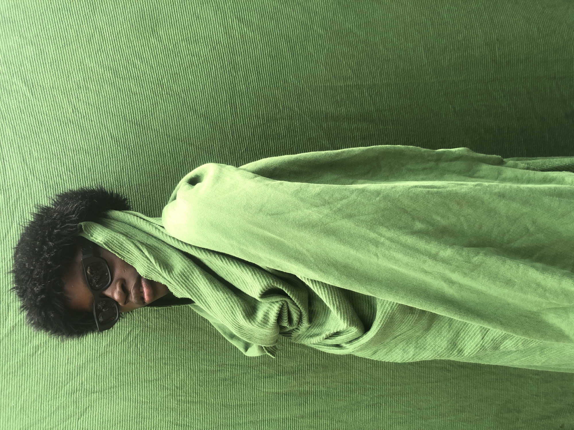 The image shows a person wearing dark sunglasses and a black fuzzy hat, draped in a vibrant green fabric, standing against a textured green backdrop. The fabric is wrapped around the person's head and shoulders, creating a draped, hooded effect. The person's face is partially visible, with their gaze directed towards the left side of the frame. The backdrop is a solid green, with a subtle horizontal ribbed texture. The lighting appears to be natural, casting soft shadows. There is no information in the image to suggest its location in Ado, Nigeria.