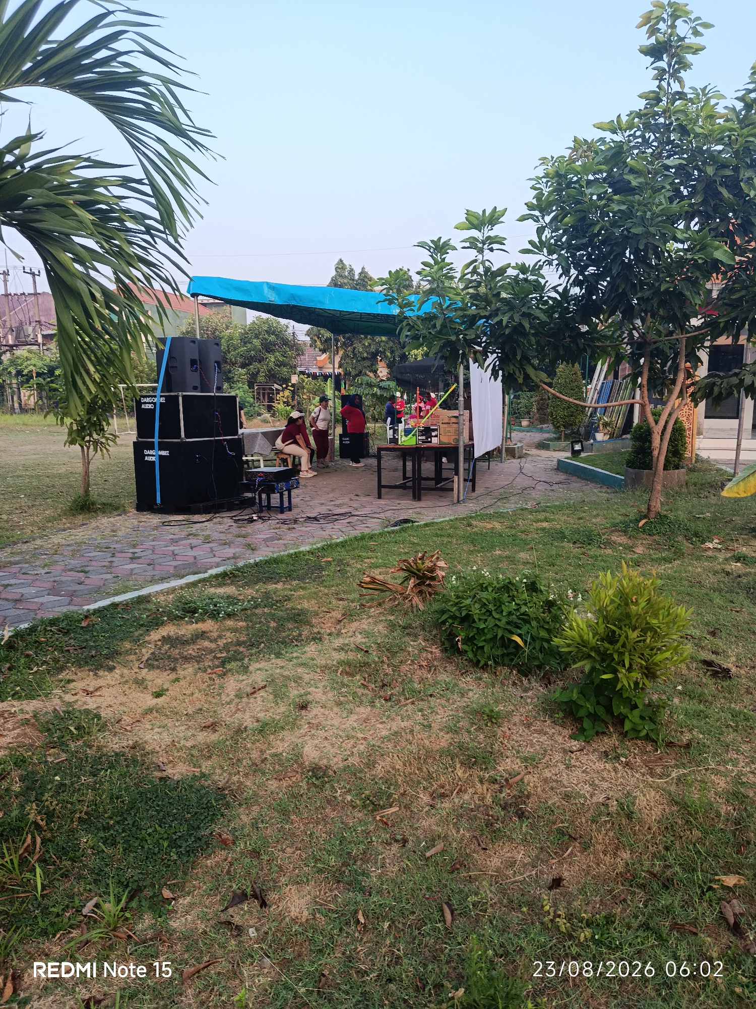The image shows an outdoor gathering or event setup in Keboansikep, Indonesia. In the foreground, there is a paved walkway made of interlocking bricks, bordered by grass. To the left, a stack of three large black audio speakers, branded "DARGO BEL AUDIO", is prominently placed. A woman in a maroon shirt and pants is seated on a small stool to the right of the speakers, facing a table. Two other individuals, one wearing a white shirt and the other a red shirt, are standing near a table under a blue tarp.
The blue tarp, supported by poles, covers a section of the area, likely serving as a makeshift stage or shelter. On the table, there are various items including what appears to be a box labeled "CASS" and some electronic equipment. To the right, a tree with lush green leaves shades part of the scene. Beyond the setup, there are more trees, buildings with tiled roofs, and power lines visible against a clear sky. The overall environment suggests a casual outdoor event in a semi-urban or suburban setting. The timestamp on the bottom right corner indicates the photo was taken on 23/08/2026 at 06:02.