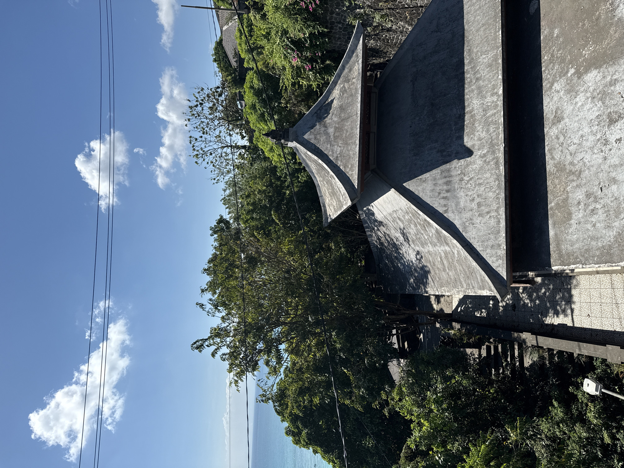 The image displays an outdoor scene from an elevated perspective in Kuta Selatan, Indonesia. The primary subject is a building with a distinctive, multi-faceted grey roof, possibly made of concrete, characterized by sharp angles and prominent shadows.
Surrounding the building is an abundance of lush green trees and dense foliage, indicating a tropical environment. Beyond the verdant landscape, a calm, blue sea is visible, extending towards the horizon. The sky is bright blue with scattered white, fluffy clouds. Several dark utility lines or power cables stretch across the upper left quadrant of the sky.
Strong sunlight illuminates the scene, casting well-defined, dark shadows from the building and trees, suggesting it is daytime, likely late morning or early afternoon. A paved or tiled area is visible near the building, partially covered by shadows. No people or specific activities are depicted in the image. The overall impression is one of a tranquil, natural setting with striking architectural elements. No visible text is present.