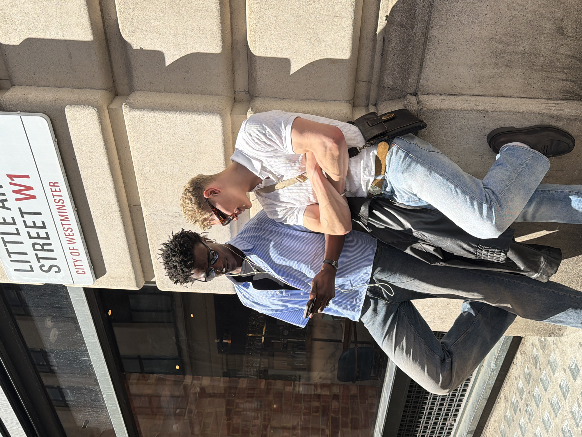 Two young men are depicted standing casually outdoors, leaning against a building facade under bright sunlight. The image is captured at a slightly Dutch angle.
The man on the left has curly blonde hair, wears dark sunglasses, a white short-sleeved polo shirt with subtle stripes, light blue jeans, and dark brown sandals. A small, dark brown or black shoulder bag with a gold clasp hangs from his left shoulder. His arms are bent, suggesting he might be holding something or simply resting them.
The man on the right has dark, curly hair, wears dark sunglasses, a light blue striped long-sleeved button-down shirt unbuttoned over a dark undershirt, and dark grey jeans. He is looking down at a black smartphone held in his right hand, with white wired earphones visible around his neck. A dark watch is on his left wrist.
Behind the subjects is a light-colored, possibly stone or concrete, building with architectural details that cast prominent shadows, indicating strong sunlight. Below the men, a large window reflects elements of the surroundings, including what appears to be a brick wall or building interior.
A white street sign is prominently visible on the left side, clearly displaying the text "LITTLE ARI STREET W1" and "CITY OF WESTMINSTER," identifying the location as London, United Kingdom. The bright light and clear shadows suggest it is daytime, likely a sunny morning or afternoon. The overall atmosphere is relaxed and casual.