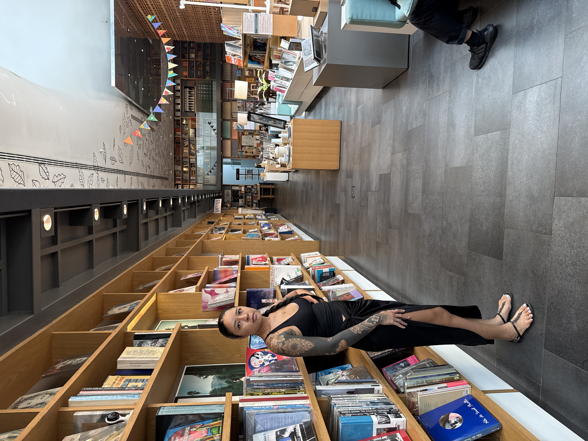 The photo captures a woman lying amidst bookshelves in a store, possibly a bookstore. She is wearing a black dress and sandals, and her arms are heavily tattooed. The woman is positioned horizontally on the edge of a shelf filled with books. The setting is a bookstore, characterized by tall, wooden bookshelves filled with a variety of books. A person wearing a black shirt and black shoes is seated in the corner of the picture. The interior of the store has a modern aesthetic with a grey tiled floor. Based on the presence of the Velaa sign, the picture was taken inside the Open House Bookstore in Bangkok, Thailand.