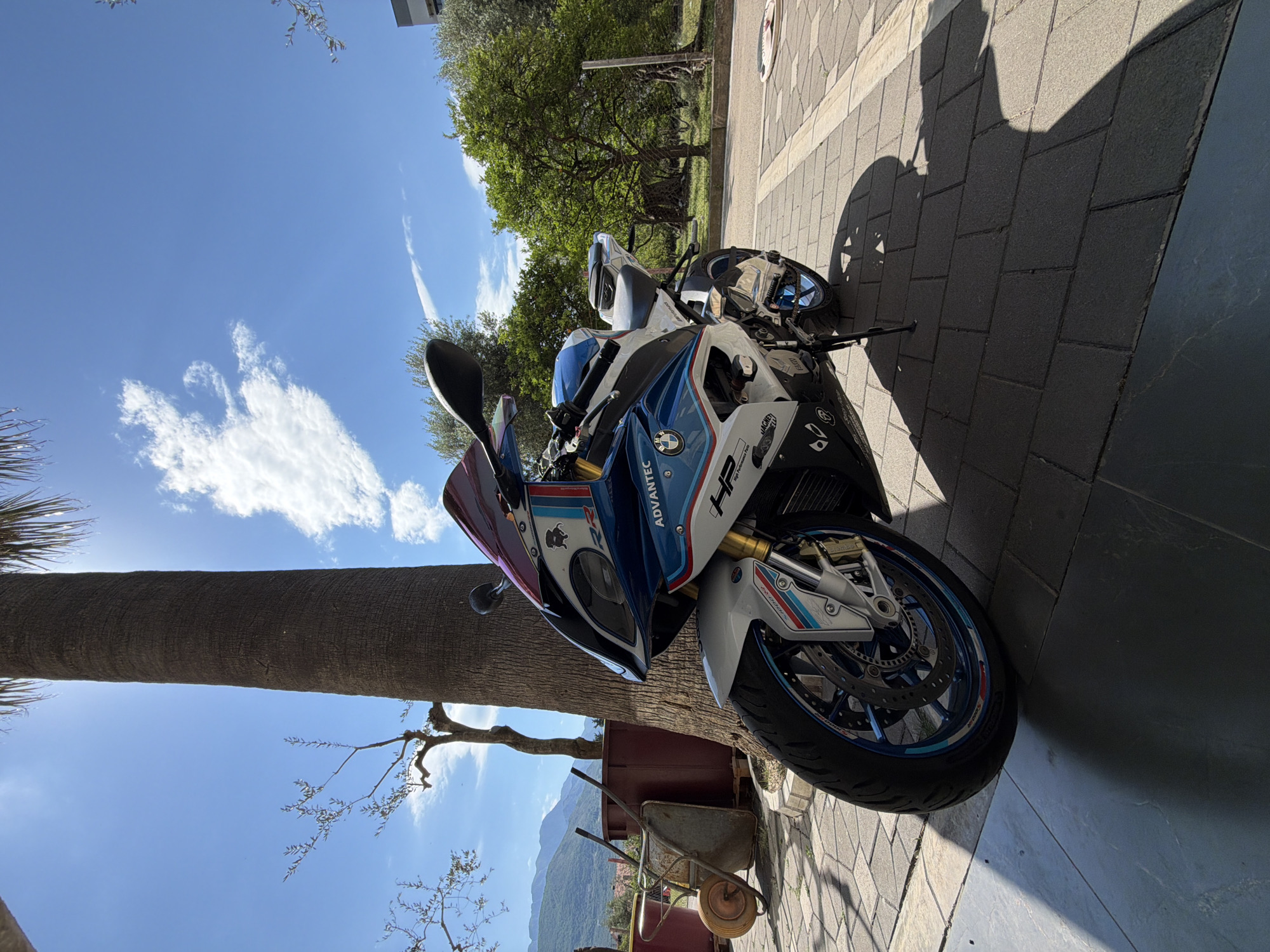 The image shows a BMW HP4 Race motorcycle parked on a paved surface, likely a sidewalk or patio, in Bar, Montenegro. The motorcycle is mostly white, blue, and red, with "ADVATEC" and "HP" logos visible. It's positioned near a thick palm tree trunk and a wheelbarrow. The background includes trees, buildings, mountains, and a blue sky with clouds. The image is taken from a low angle and rotated. The shadow of the motorcycle is cast on the pavement. The natural environment and architecture are consistent with the coastal setting of Bar. No people are visible.