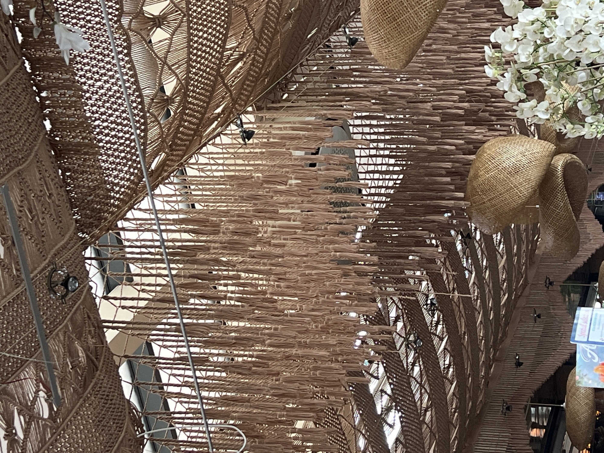 The image displays an elaborate indoor decorative ceiling, likely within a shopping mall or large public building in Varna, Bulgaria. The overhead installation is composed primarily of natural fibers in various shades of brown and beige, creating a warm, organic aesthetic.
Dominating the scene are large, curved structures crafted from intricately woven or braided natural materials, resembling macrame or a complex basket weave. These sections feature diverse patterns, including geometric designs and open meshwork, through which the bright ceiling and structural elements of the building are visible. Adjacent to these woven sections are areas covered with densely packed, short bundles of fiber, forming a textured, fringed surface that adds depth to the installation.
Several decorative objects, made from what appears to be burlap or a similar coarse fabric, are suspended throughout the display. These include spherical forms and larger, bow-like shapes. A prominent cluster of artificial white flowers, possibly orchids, hangs towards the upper right, providing a delicate floral accent against the natural tones. Small, dark lighting fixtures are subtly integrated into the fiber structures, illuminating the space.
In the bottom right corner, partially visible are colorful tags with faint text, including "Special selection" and a brand name that appears to be "Aloe" or similar, alongside a floral illustration. This detail further suggests a commercial or retail setting. The overall design creates a unique and inviting atmosphere, blending natural materials with a sophisticated, artful arrangement.