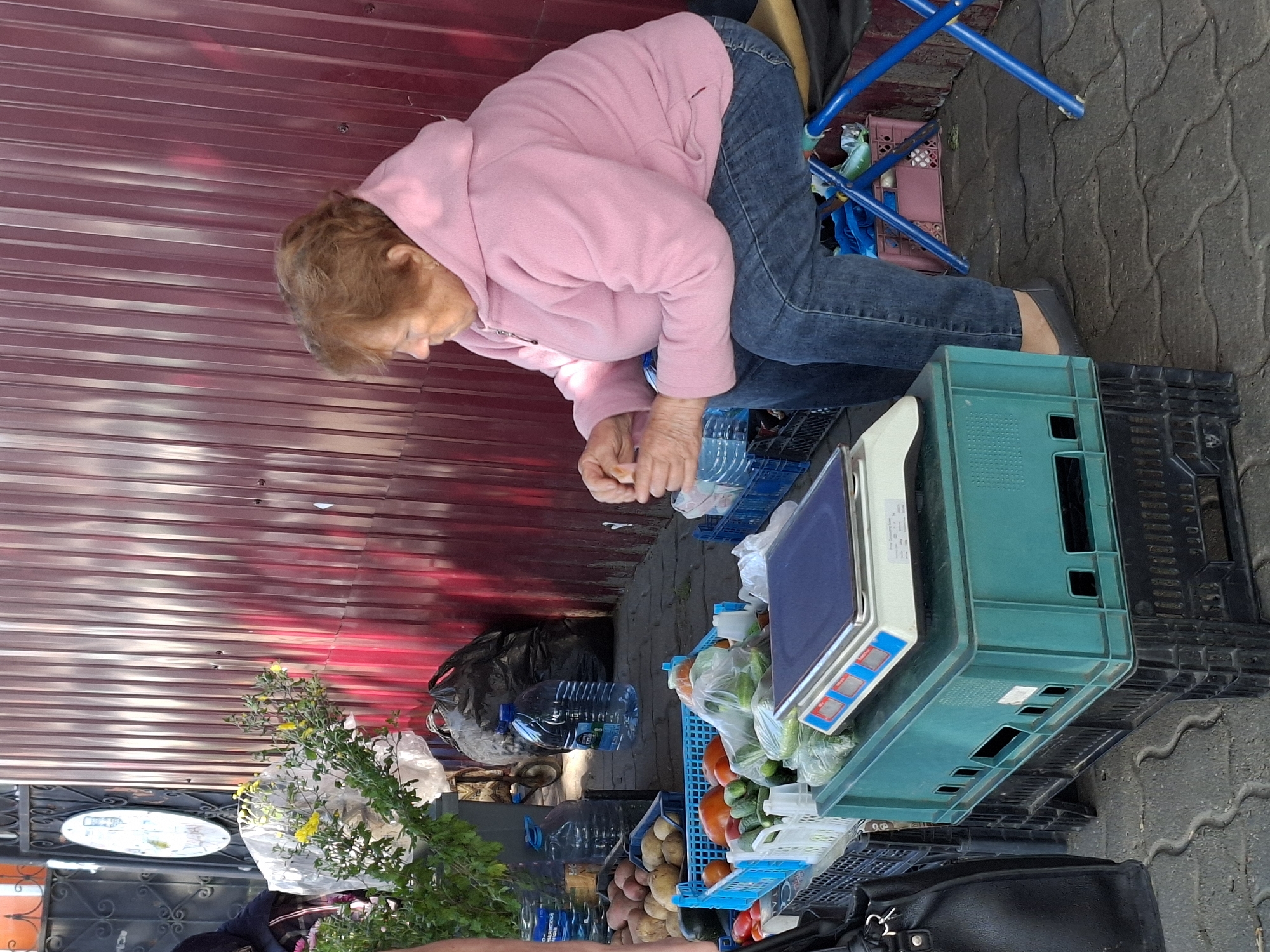 An elderly woman with auburn hair, wearing a pink hooded jacket and blue jeans, sits on a stool in front of a stall displaying fresh produce. She is holding a small item in her hands, seemingly examining it. The stall features various vegetables, including tomatoes, cucumbers, and potatoes, arranged in blue crates. A digital scale sits on top of a teal and black plastic crate, ready for weighing purchases. Behind the stall, a large black garbage bag and several large bottles of water are visible, leaning against a corrugated metal fence. The scene appears to be an outdoor market, with a textured stone pavement underfoot. The overall environment is bright and appears to be daytime.