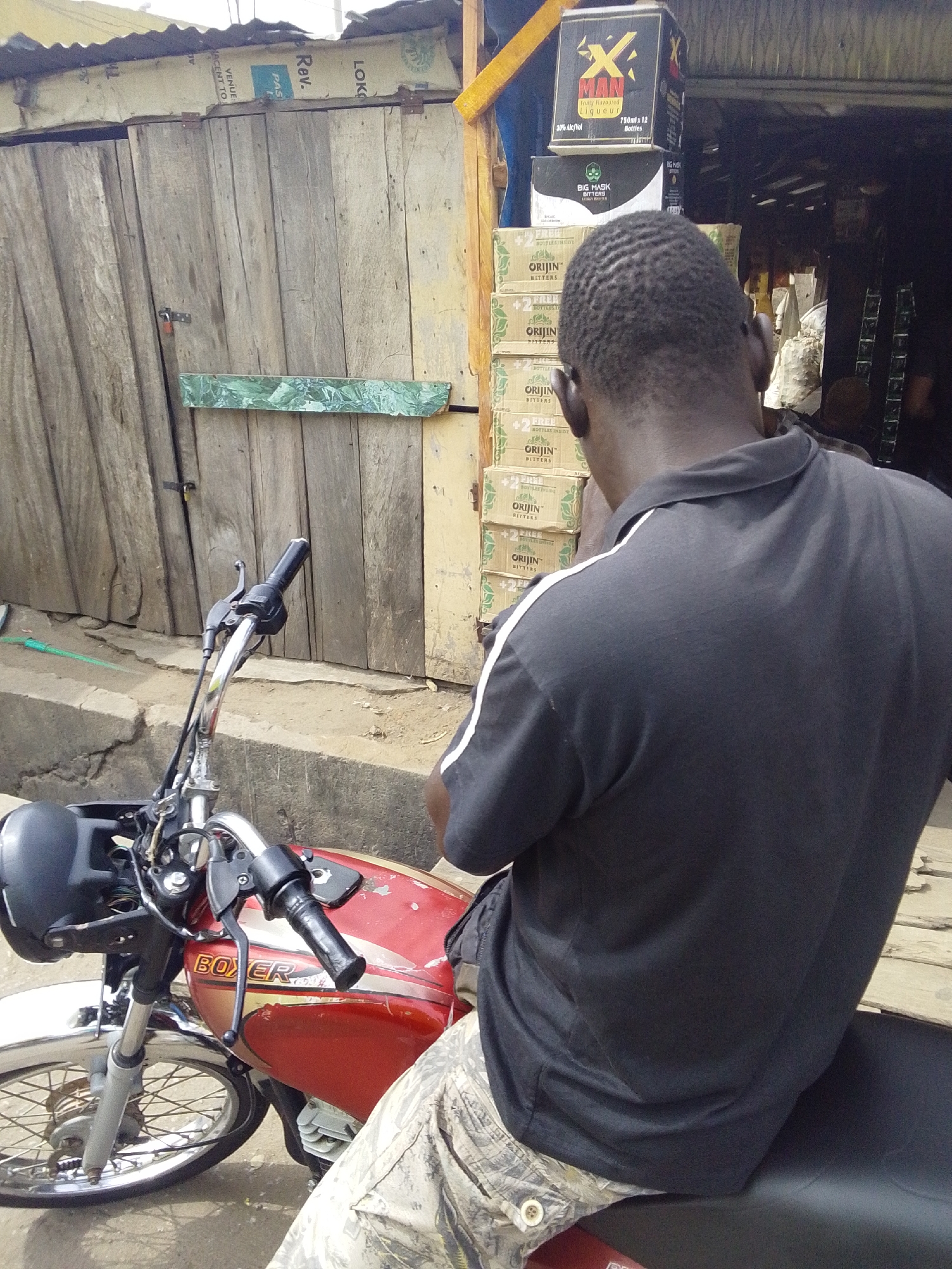 A man wearing a black polo shirt with white stripes on the shoulders and camouflage pants is sitting on a red Boxer motorcycle. He faces away from the camera, towards a small storefront in Lokoja, Nigeria. The storefront has a wooden facade and displays stacked boxes of "ORIJIN Bitters," "BIG MASK Bitters," and a box of "X MAN Fruity Flavoured Liqueur."