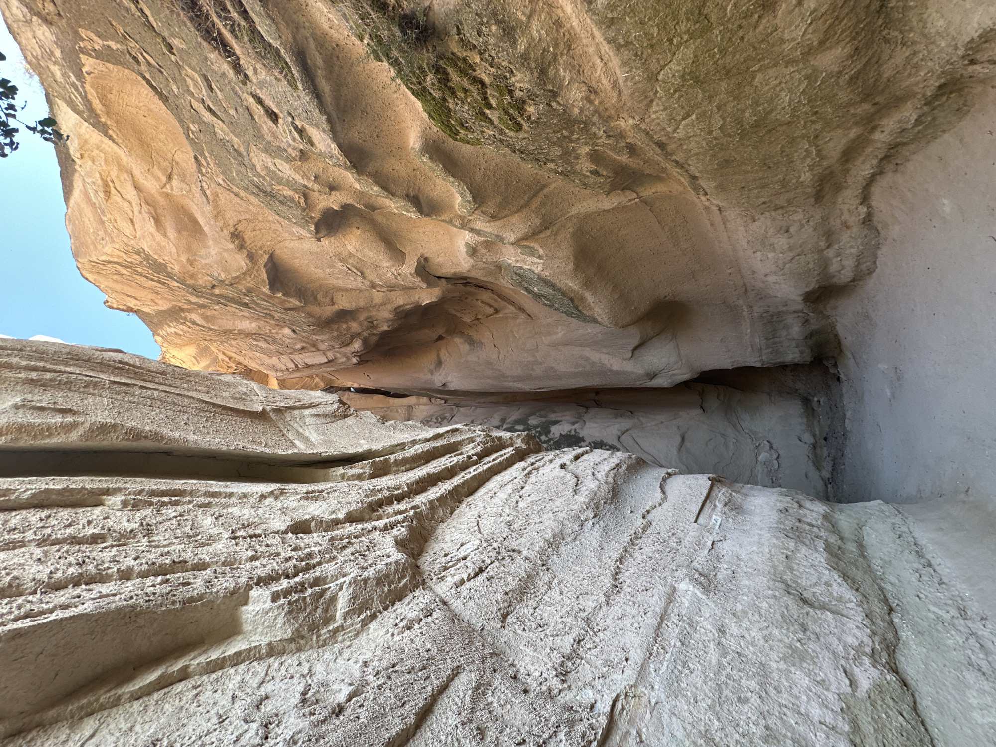The image shows a narrow canyon with towering, sculpted rock walls. The left wall is a pale, textured stone with prominent horizontal striations and a rough, pitted surface, suggesting erosion over time. The right wall is a warmer, sandy-colored rock, also showing signs of weathering with smooth, undulating curves and some patches of green moss. The canyon floor is a light-colored, sandy path leading into the shadowed depths. The sky above is a clear, bright blue. This scene depicts a natural geological formation, likely a result of wind and water erosion, located in the Cappadocia region near Avanos, Turkey, known for its unique "fairy chimney" rock formations and cave dwellings. There are no people or specific objects visible, emphasizing the raw, natural environment.