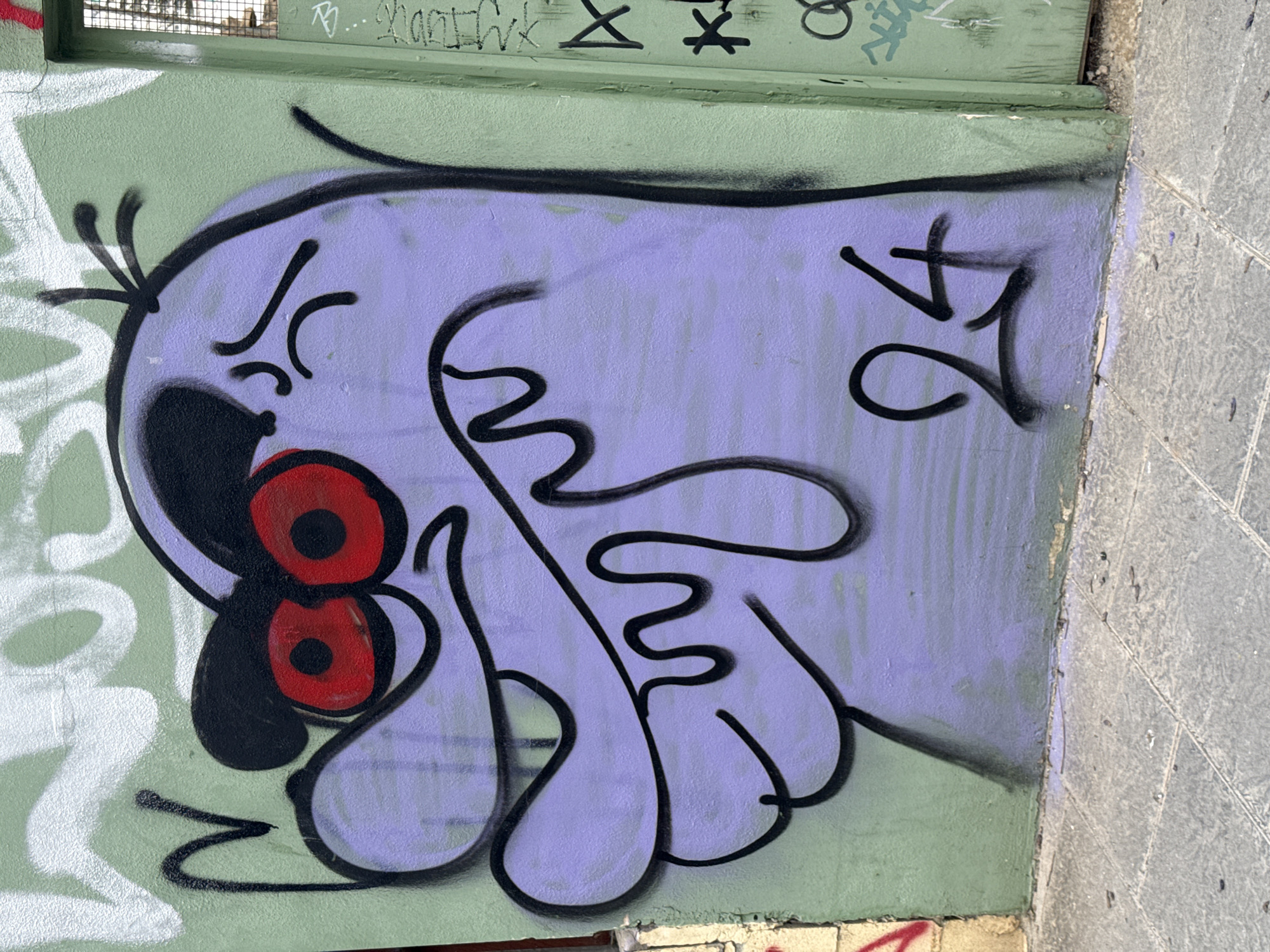 Graffiti depicting an angry cartoon character with red eyes is painted on a green wall at an unidentified location. The character is purple with black outlines. The number "24" is painted on the right side. The wall is also covered in other graffiti. A sidewalk is visible to the right.