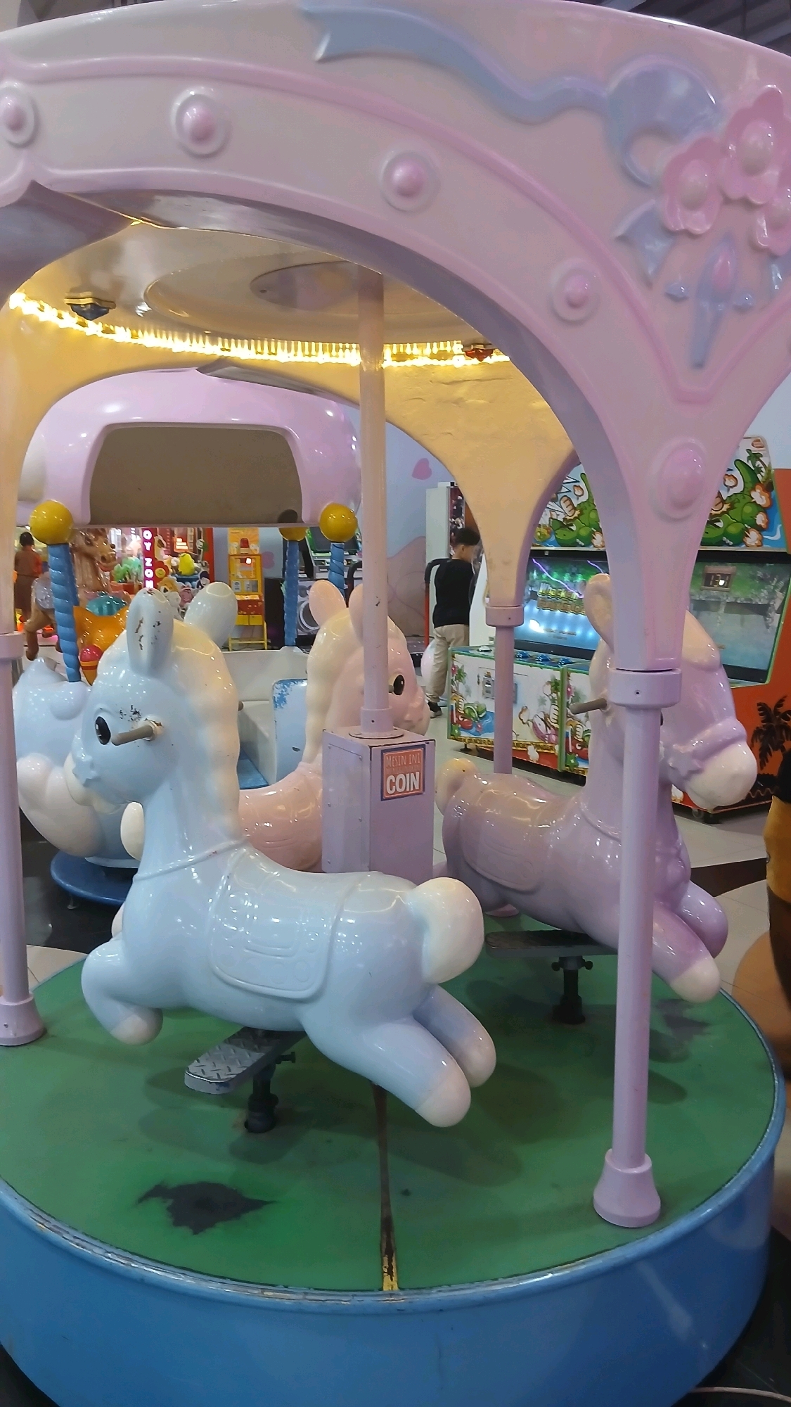 This image captures a colorful carousel with plastic horses in a public indoor space, likely a mall or arcade, in the city of Binjai, Indonesia. The carousel's canopy is adorned with pastel pink and white decorative elements, including sculpted flowers and rounded buttons. Beneath the canopy, several carousel horses are visible. One horse in the foreground is a pale blue, while another to its right is light pink. Further back, a pale purple horse is partially visible. The horses are mounted on poles and appear to be stationary. A coin slot mechanism is attached to a purple post between two of the horses, with a sign reading "MAIN INI COIN" which translates to "PLAY THIS COIN". The carousel floor is a vibrant green, marked with lines, and a blue base encircles the entire structure. In the background, arcade game machines with bright lights and colorful graphics are present, along with a few indistinct figures of people, one of whom appears to be a child wearing a black shirt and beige pants, standing near the game machines. The lighting suggests an indoor environment with artificial illumination.