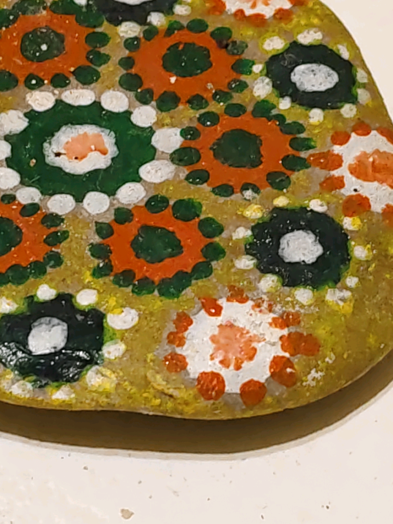 A close-up, overhead shot captures a painted stone with a yellow-green base, adorned with a pattern of dots and circular designs. The circular designs feature orange, green, and white elements, creating a floral or mandala-like motif. Some circles are filled with green and have white centers, while others are orange with green dots surrounding them. Small white and yellow dots are scattered across the stone's surface. The stone itself has an irregular, somewhat flattened shape and appears to be resting on a light-colored, speckled surface. The lighting is bright, casting a slight shadow on the bottom right side of the stone, suggesting it is positioned indoors. The image does not provide any context related to Rostov-on-Don, Russia, or any human presence.